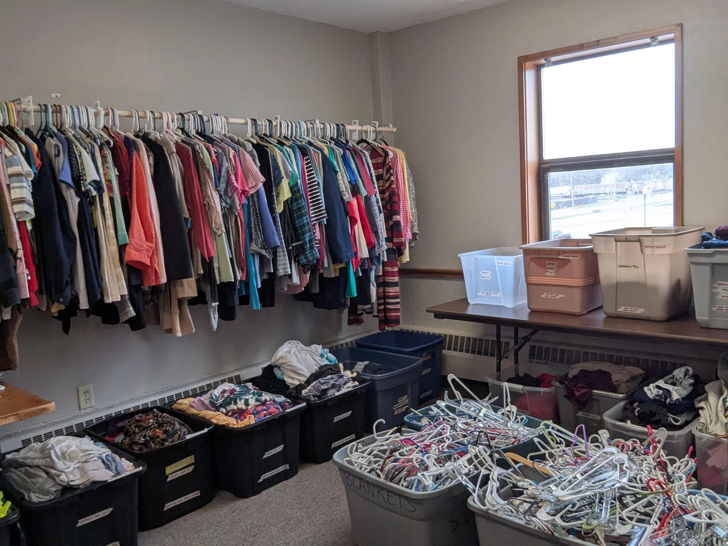 A room with clothes on hangers, plastic bins of folded clothing, and boxes of miscellaneous items, indicating a space for sorting or donations.