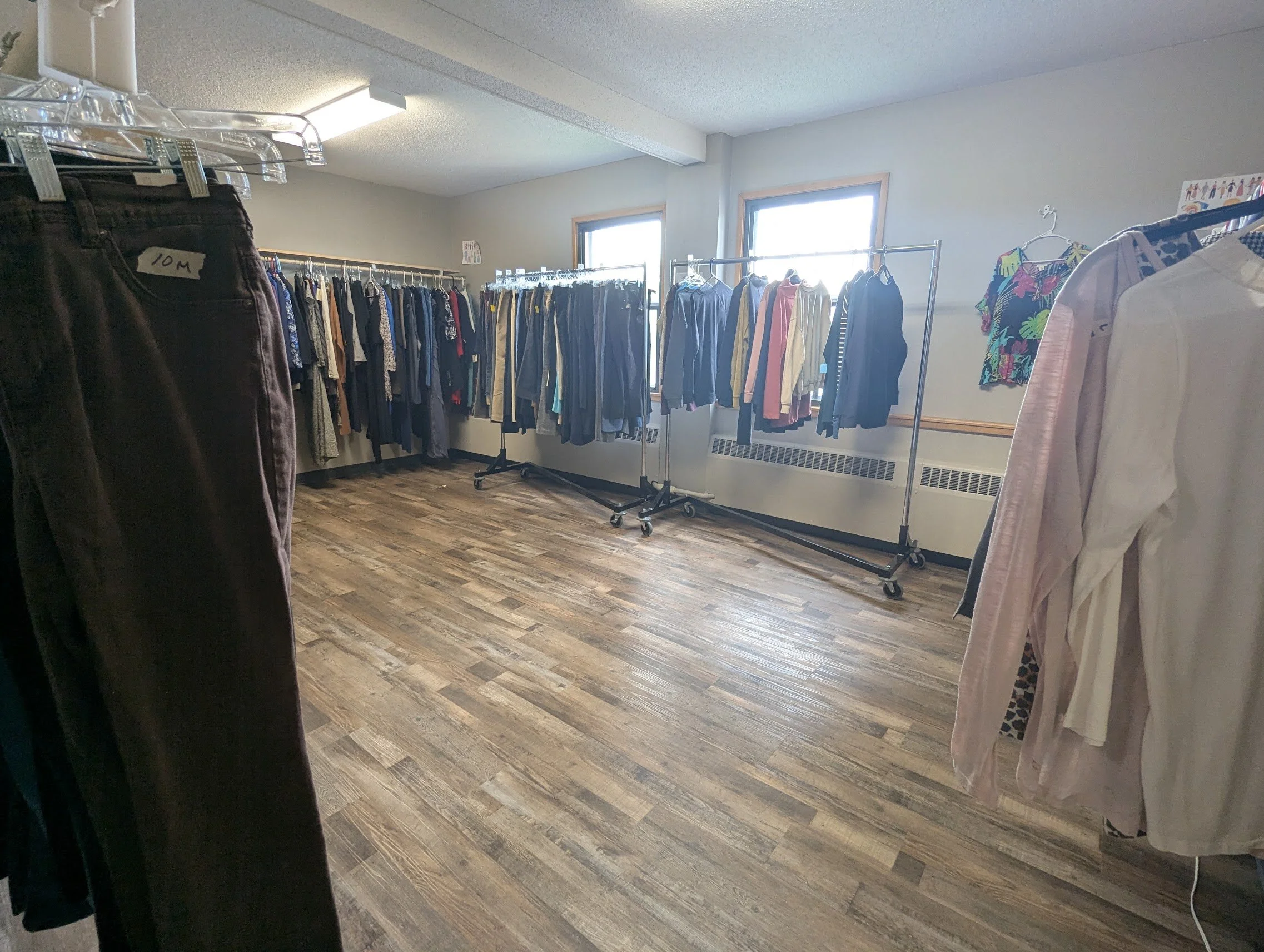 Clothing racks with various shirts, jackets, and pants in a room with wooden flooring and two windows.