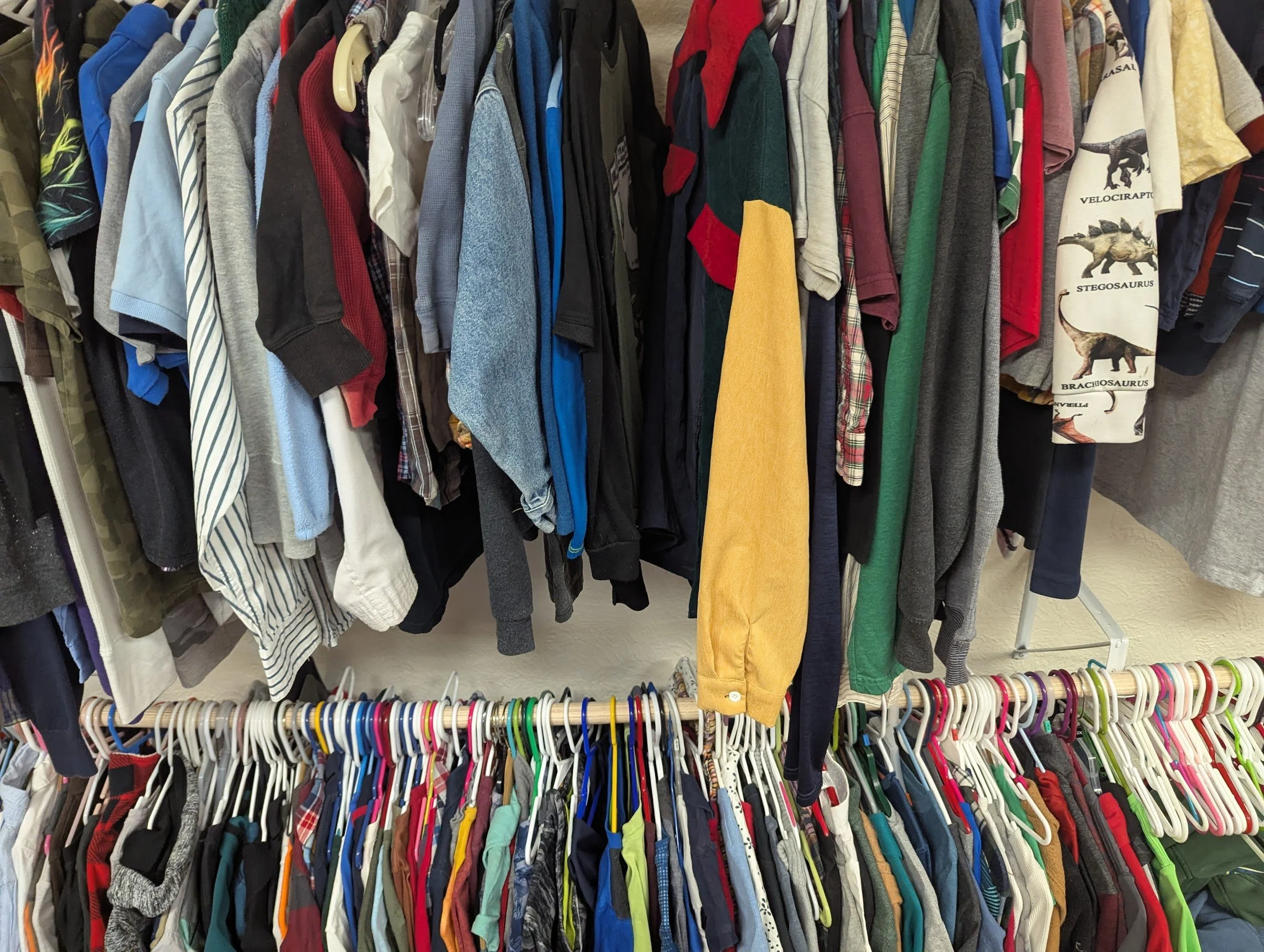 Clothing rack with various men's and boys' shirts and t-shirts hanging on plastic and wooden hangers, including polo shirts, t-shirts, and dress shirts of different colors and patterns, at a thrift store or clothing store.