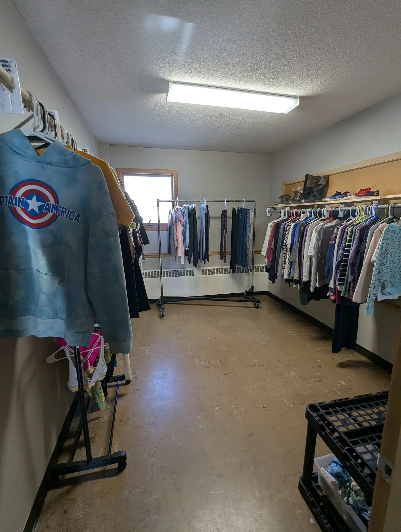 A thrift store or donation center with clothing on racks and shelves, a window letting in natural light, and a floor with a tan-colored surface.