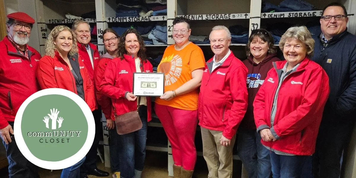 Group of nine people standing in front of shelves labeled 'Denim Storage,' with one person holding a framed check. Most are wearing red jackets or shirts, and one woman is wearing an orange t-shirt. A logo for 'Community Closet' is in the bottom left corner.