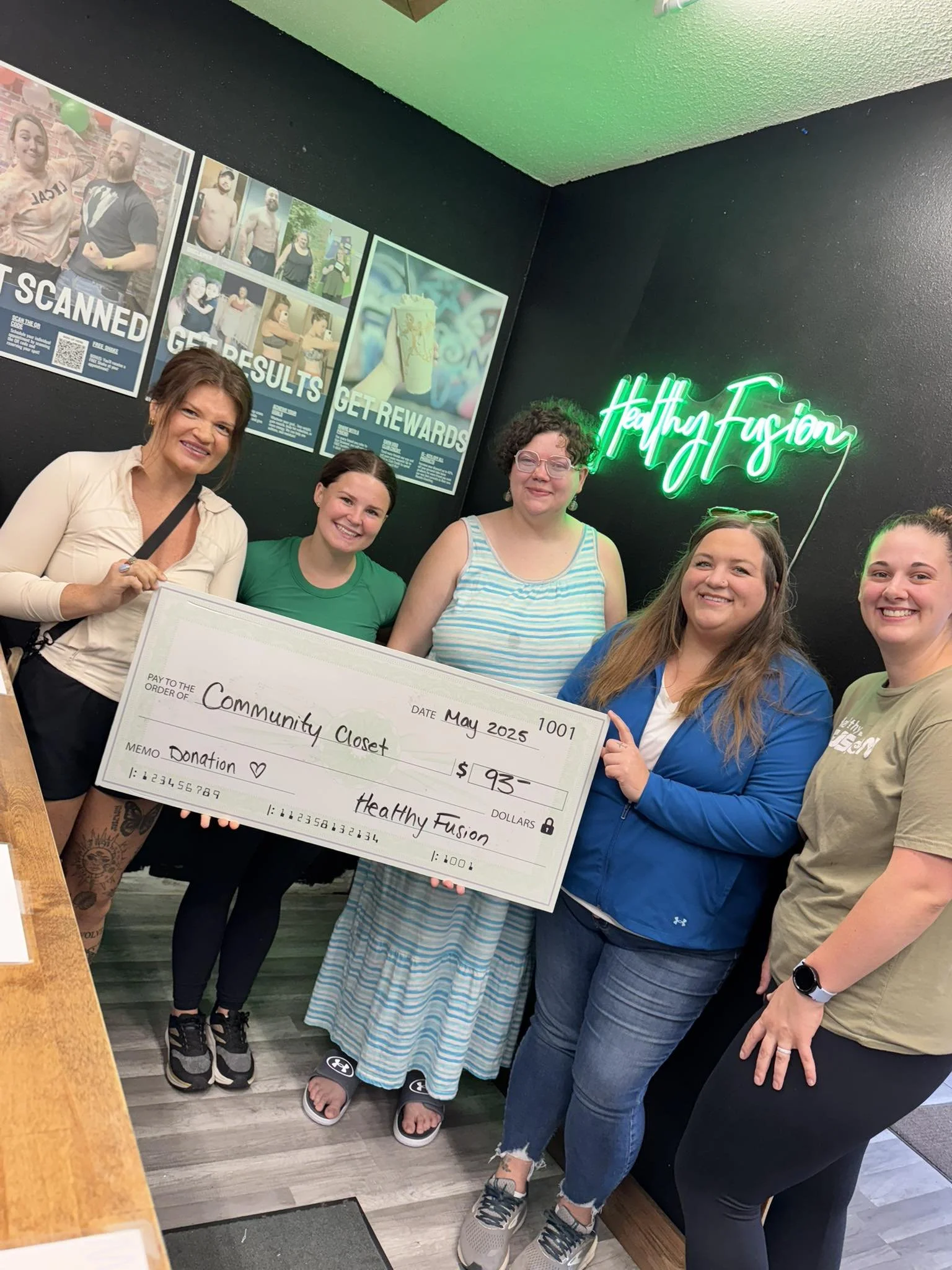 Group of five women standing indoors, holding a large check made out to Community Closet. The women are smiling, and the background has posters and a neon sign that says Health Fusion.