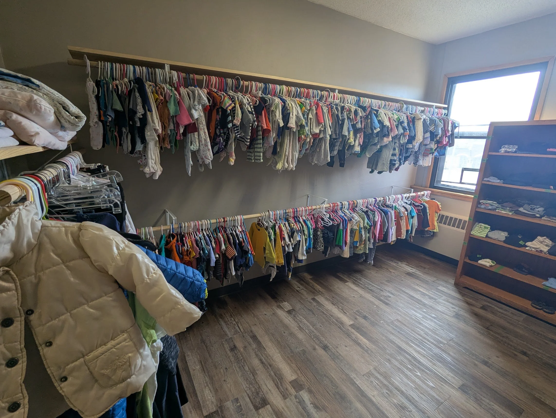 Clothing items on hangers on a wall-mounted rack, with folded clothes on a shelf on the left and additional clothes on hangers at the bottom, in a room with a window allowing natural light.