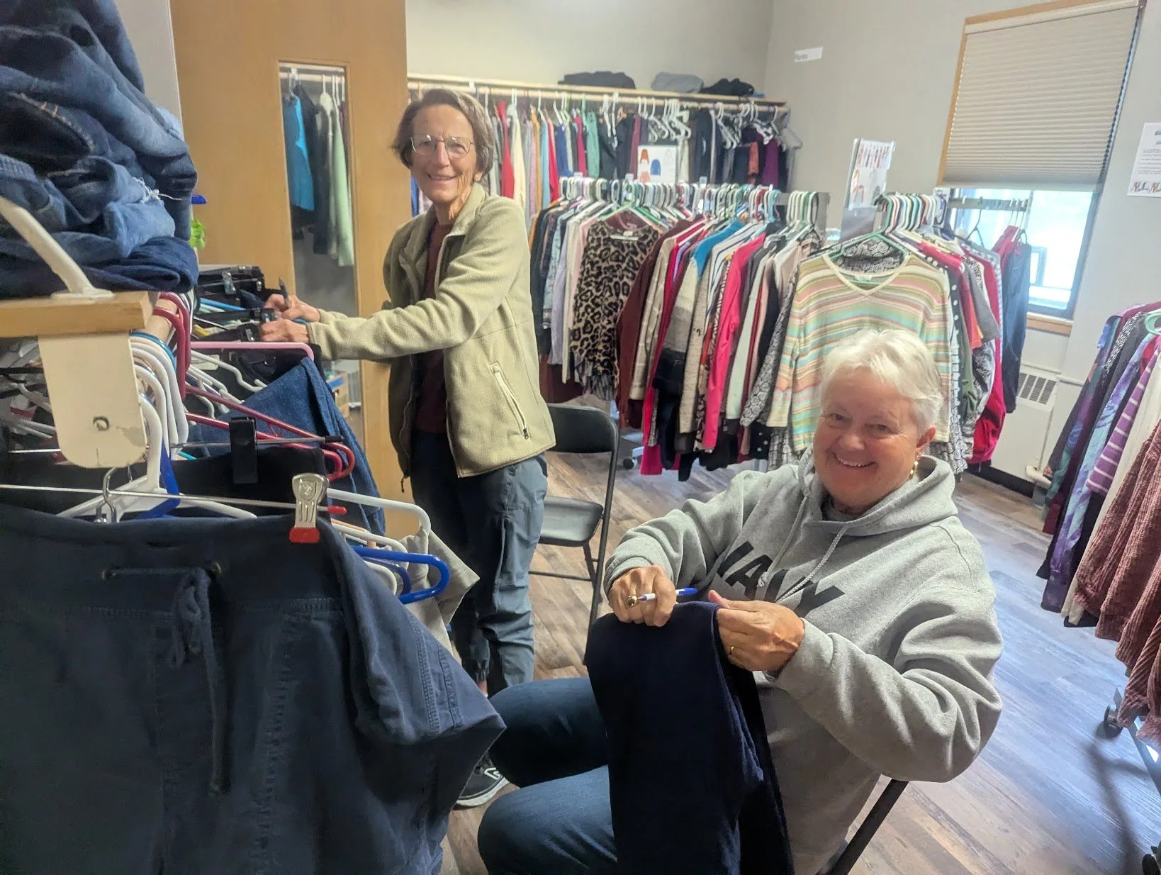 Two women shopping for clothes in a thrift store, one standing and the other sitting, both smiling. There are racks of clothing behind them.