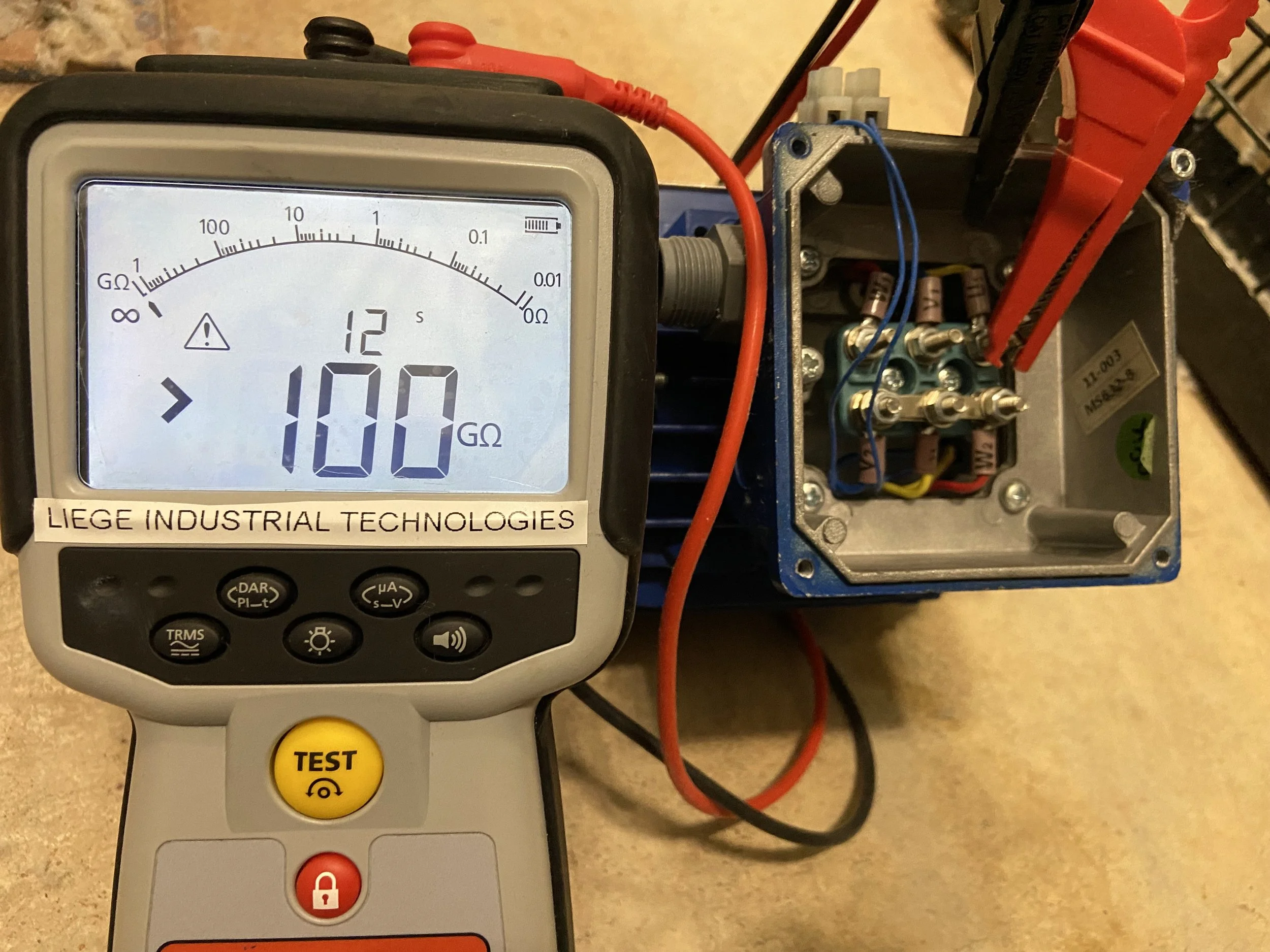 A digital multimeter displaying a resistance measurement of 100 ohms, connected to an electrical device with wires and components inside a metal enclosure.