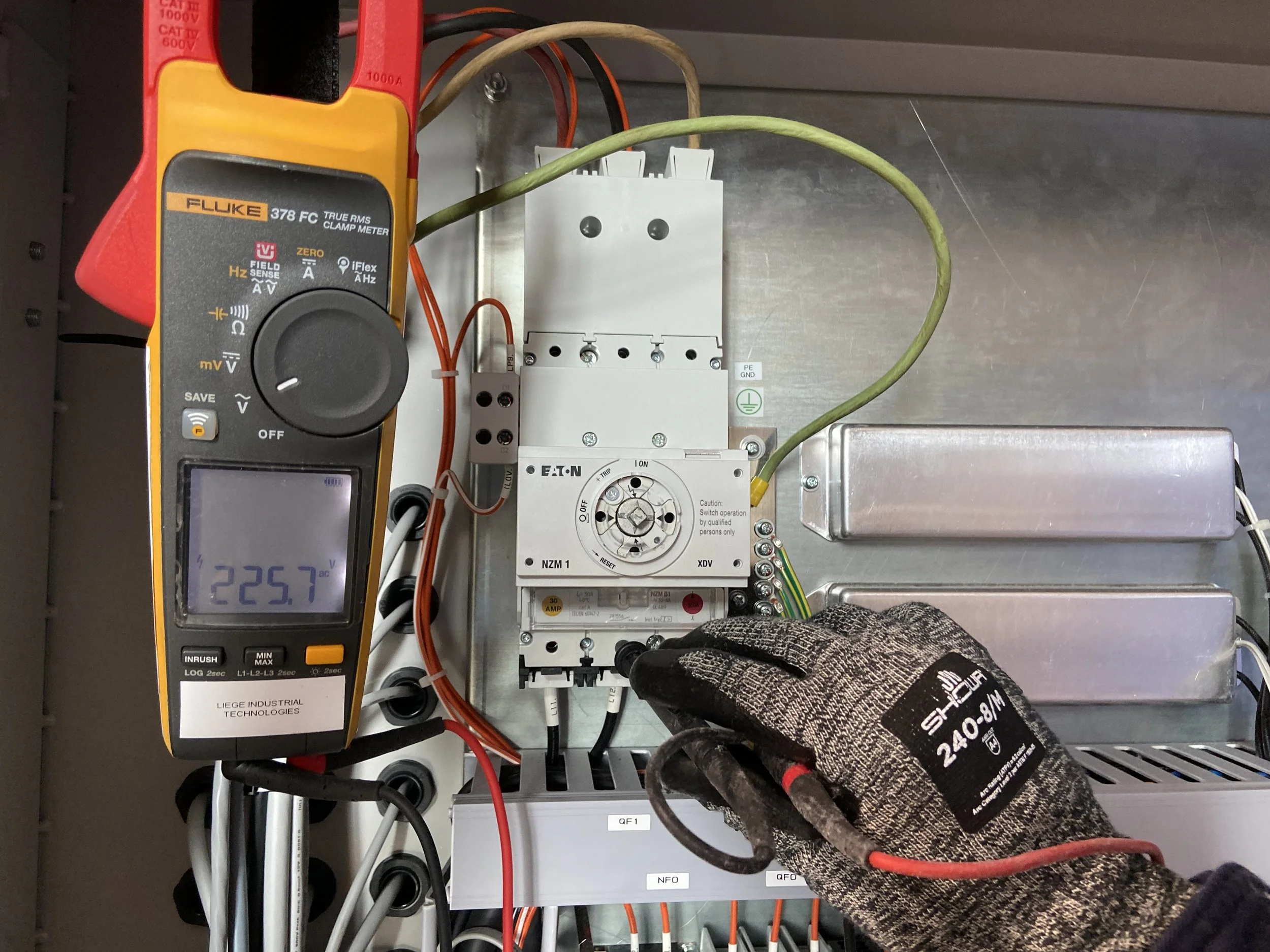 A person wearing a glove is using a multimeter to measure electrical current on a circuit breaker panel. The multimeter displays a reading of 225.7 volts.