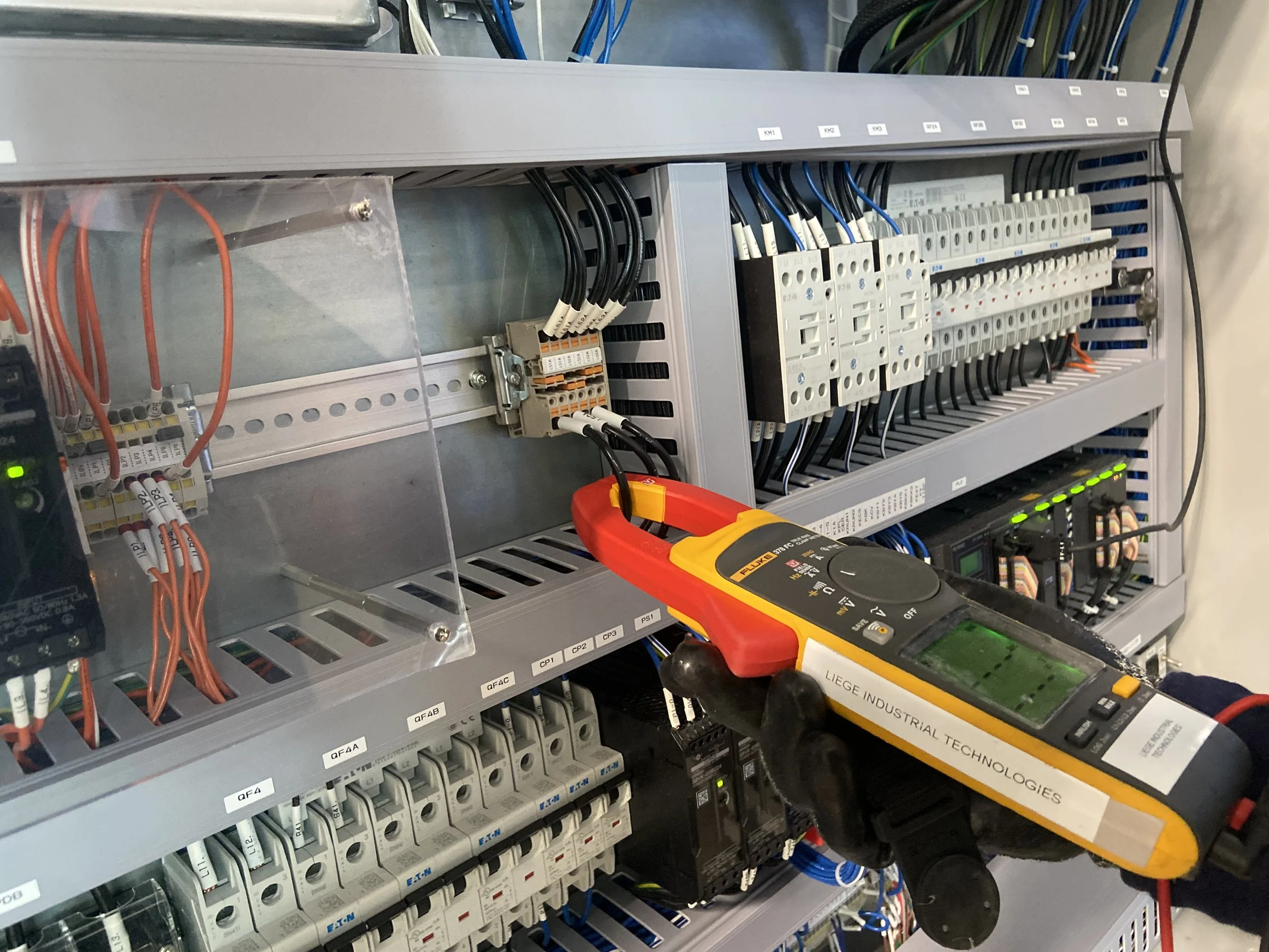 A technician uses a multimeter to test wiring inside an electrical control panel with various circuit breakers, wires, and components.