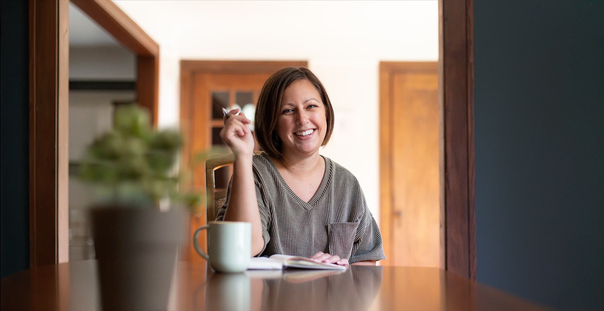 A woman in her thirties sits at a dining table with a notebook mug and pen smiling at the camera