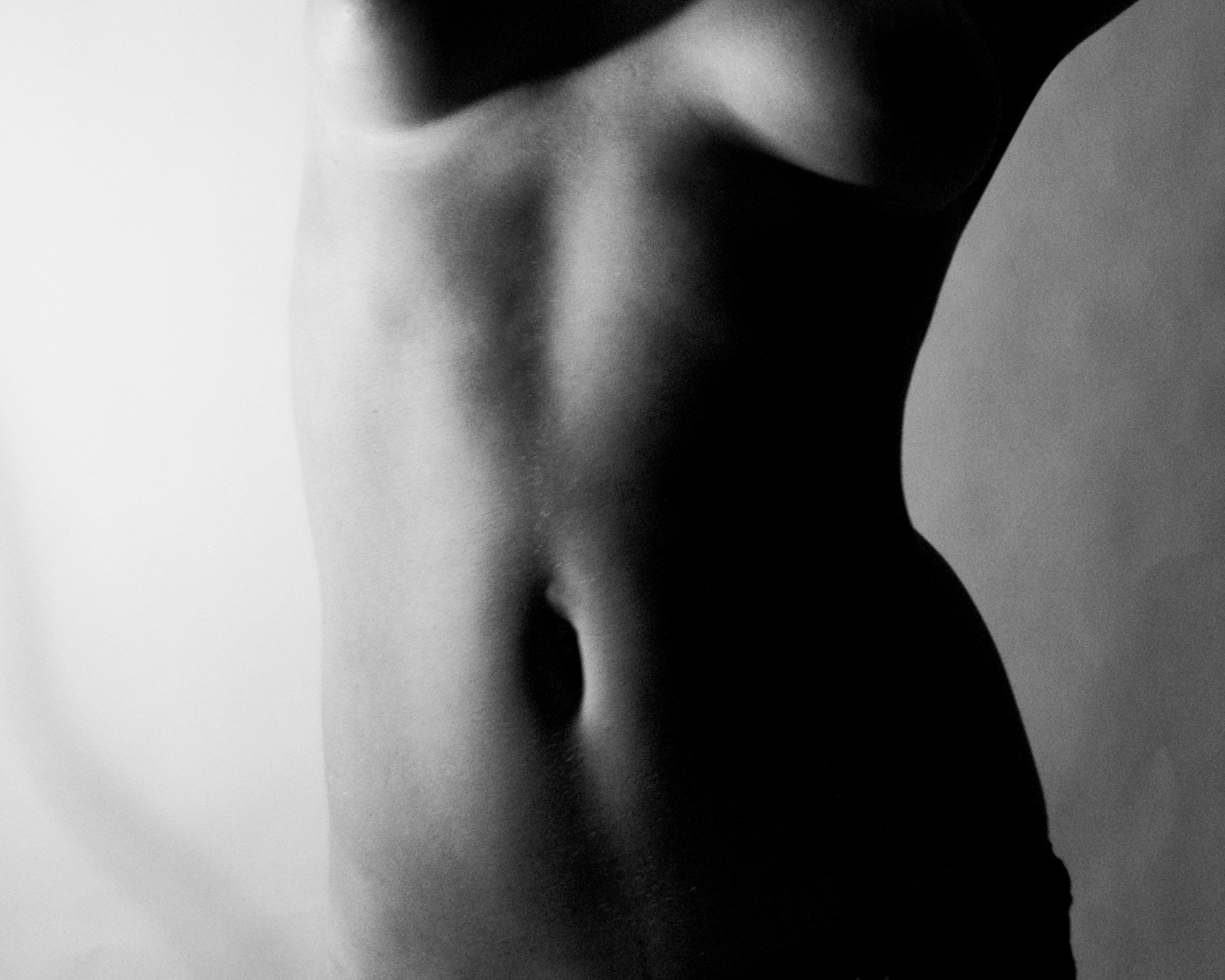 Black and white photo of a woman's torso, showing her bare midriff and side, with shadowed contours and a neutral background.