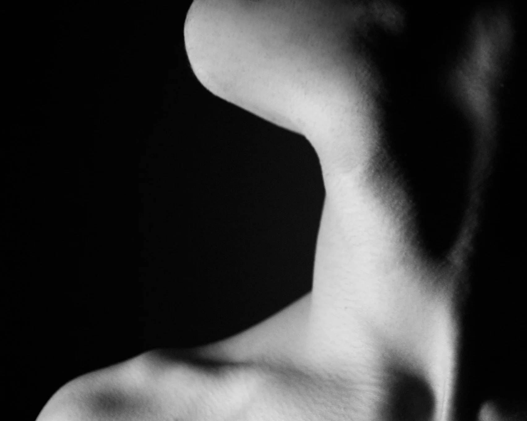 Close-up black and white photograph of a person's shoulder and neck with soft lighting, with a dark empty space obscuring the face.