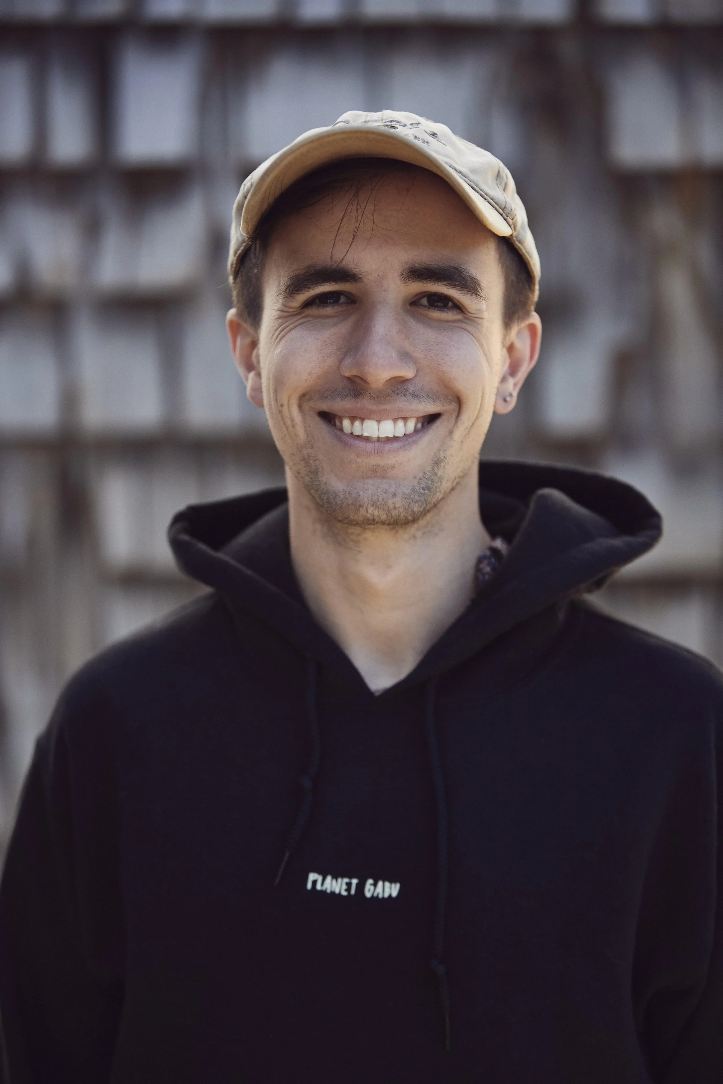 A smiling young man wearing a beige cap and black hoodie with "Planet Gadu" logo, standing outdoors in front of a wooden fence.