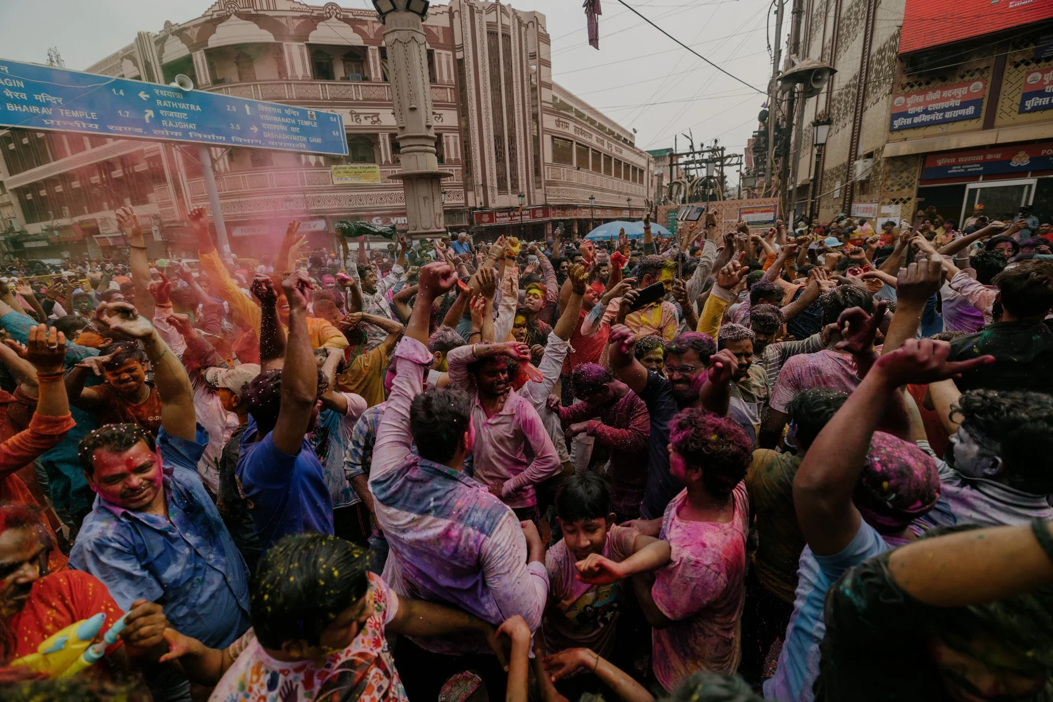 Large crowd of people celebrating holi on a street, with colorful powder on their faces and clothing, in front of buildings and signs in Hindi, during daytime.