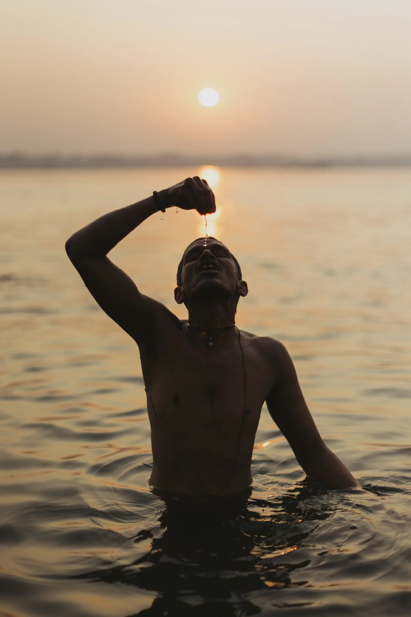 A man in the water at sunset, pouring water over his face with the sun in the background.
