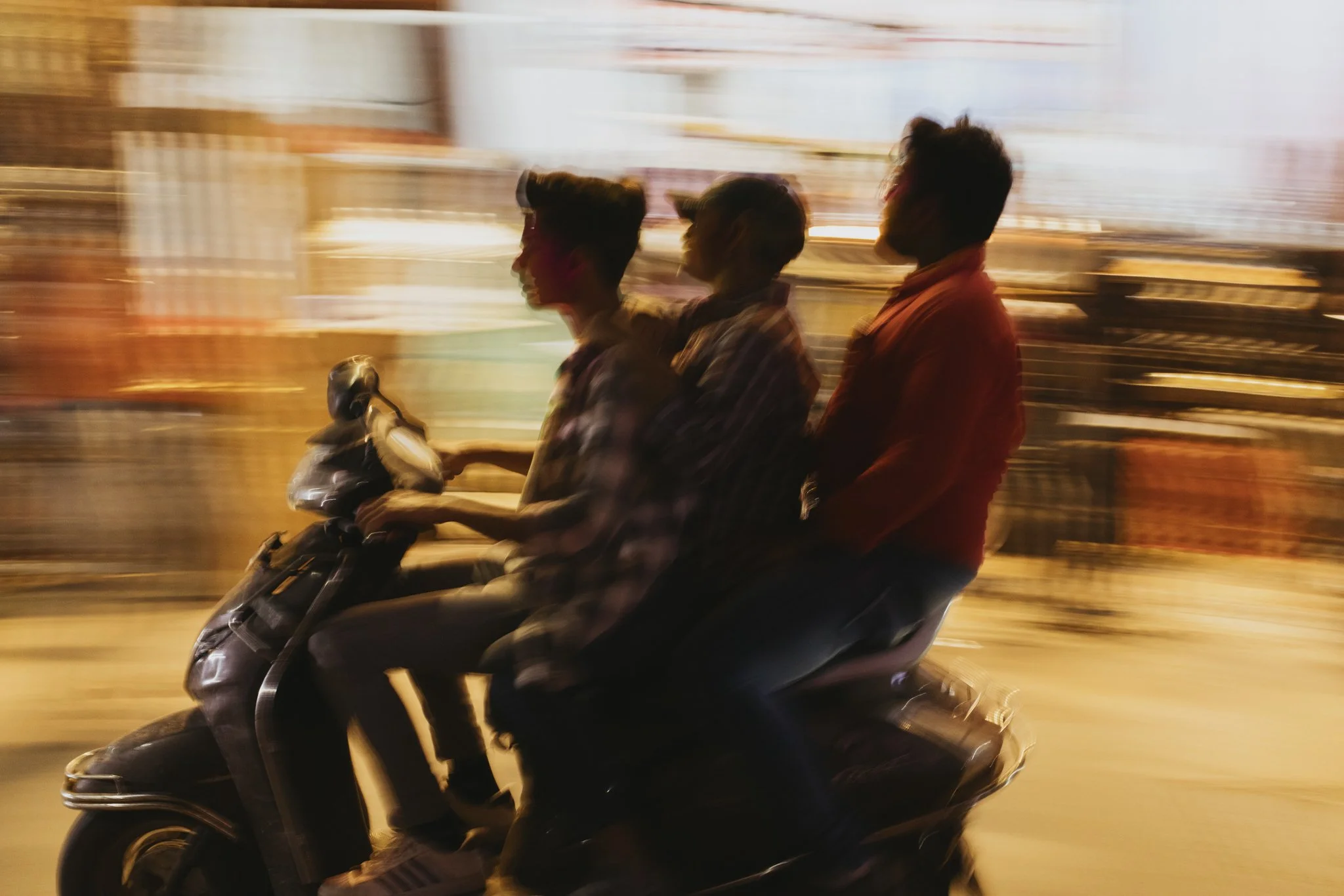 Three young men riding a scooter at night with a blurred city background.