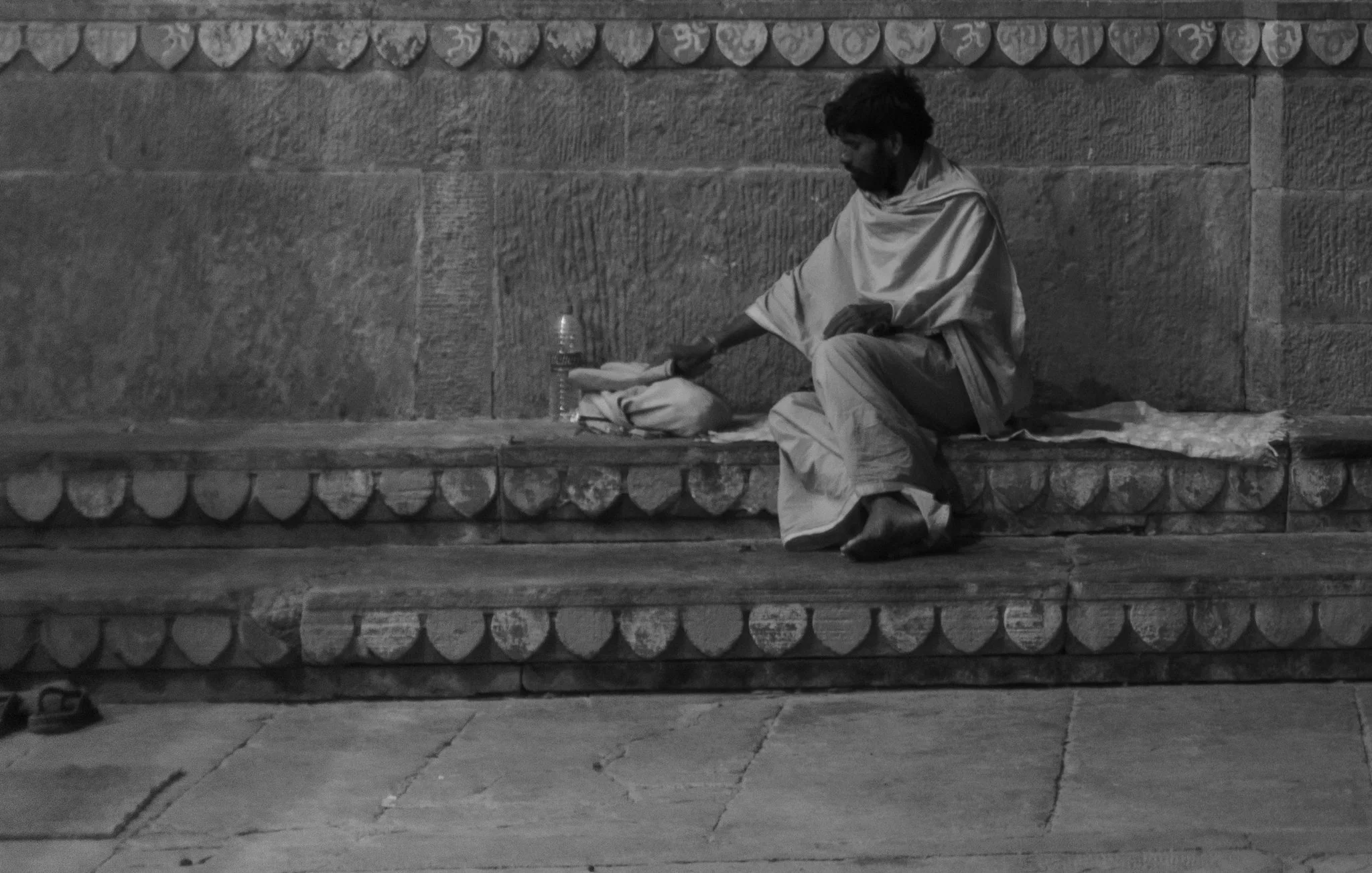 A man sitting alone on a stone bench with a water bottle and some items in front of him, against a stone wall decorated with a row of carved stones.