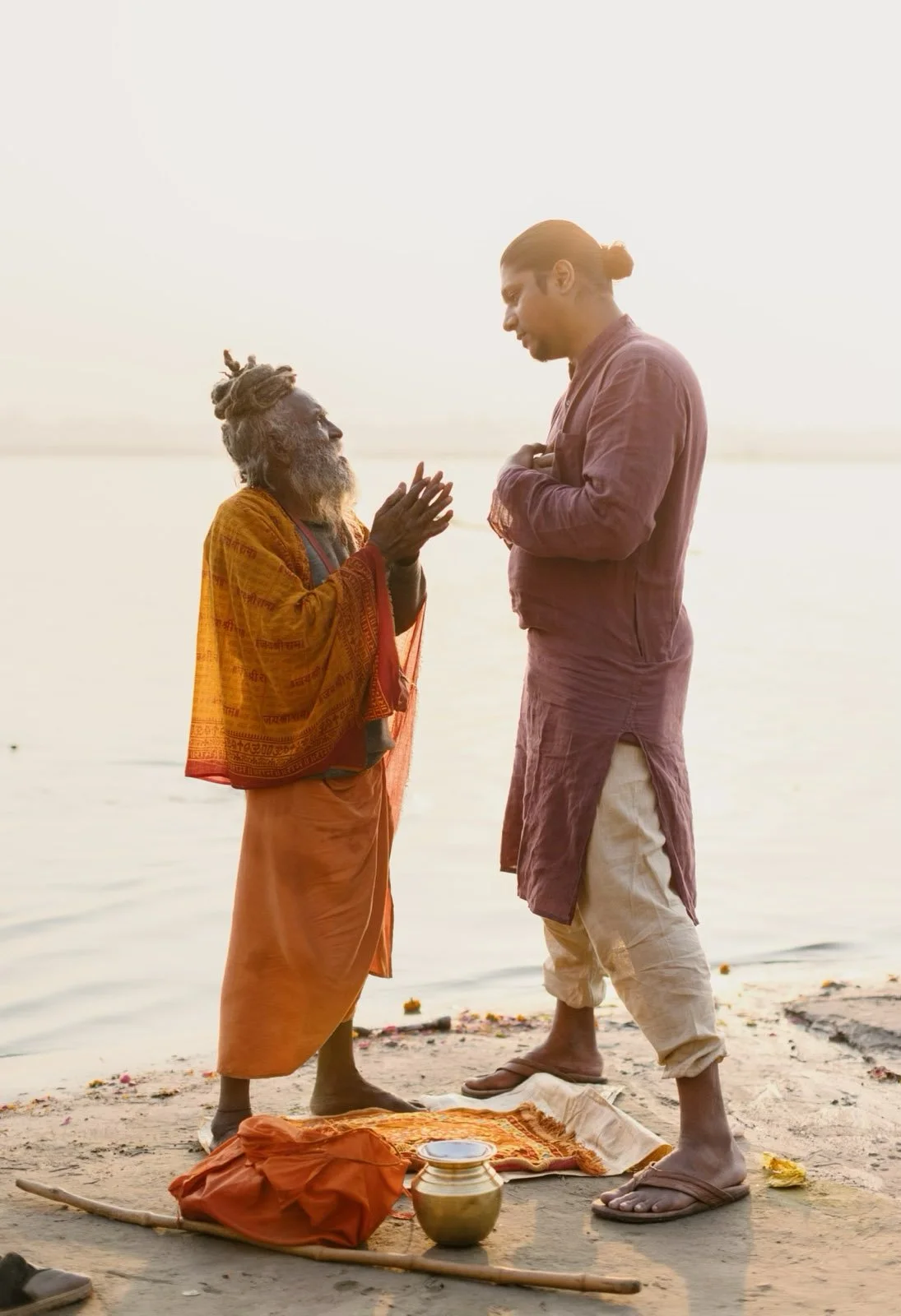 An elderly man with dreadlocks and traditional clothing praying with a younger man on a sandy shore during sunset or sunrise.