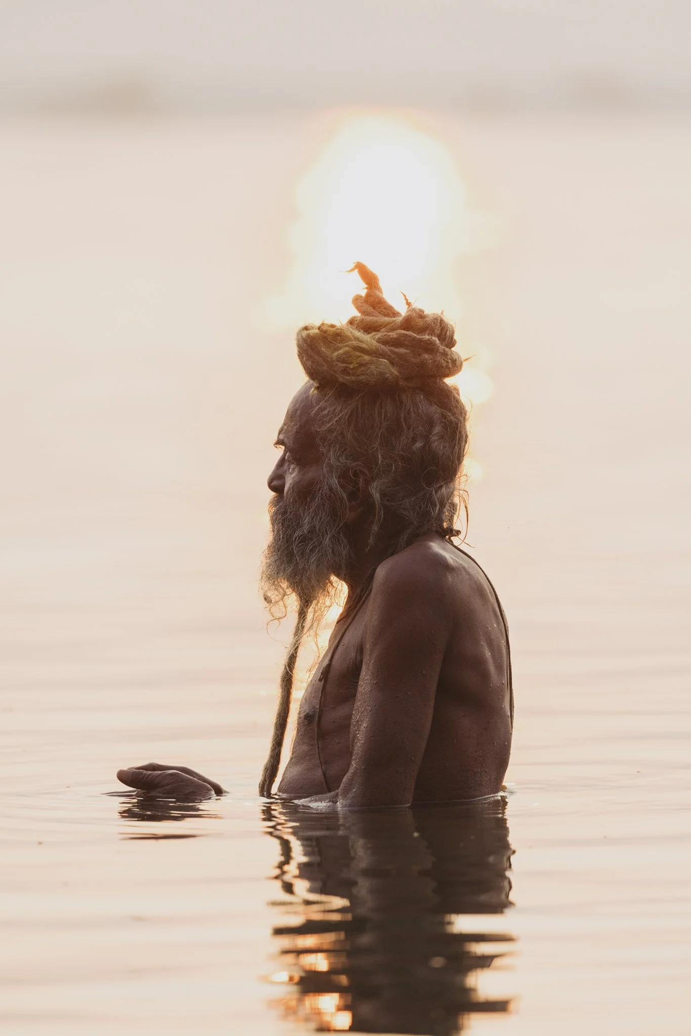 An elderly man with long dreadlocks and a beard stands in water during sunset, with the sun positioned behind his head creating a halo effect.