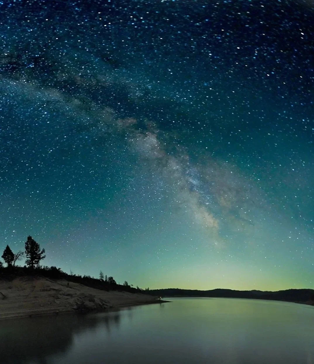 Night sky filled with stars over a body of water with a silhouetted treeline on the horizon.
