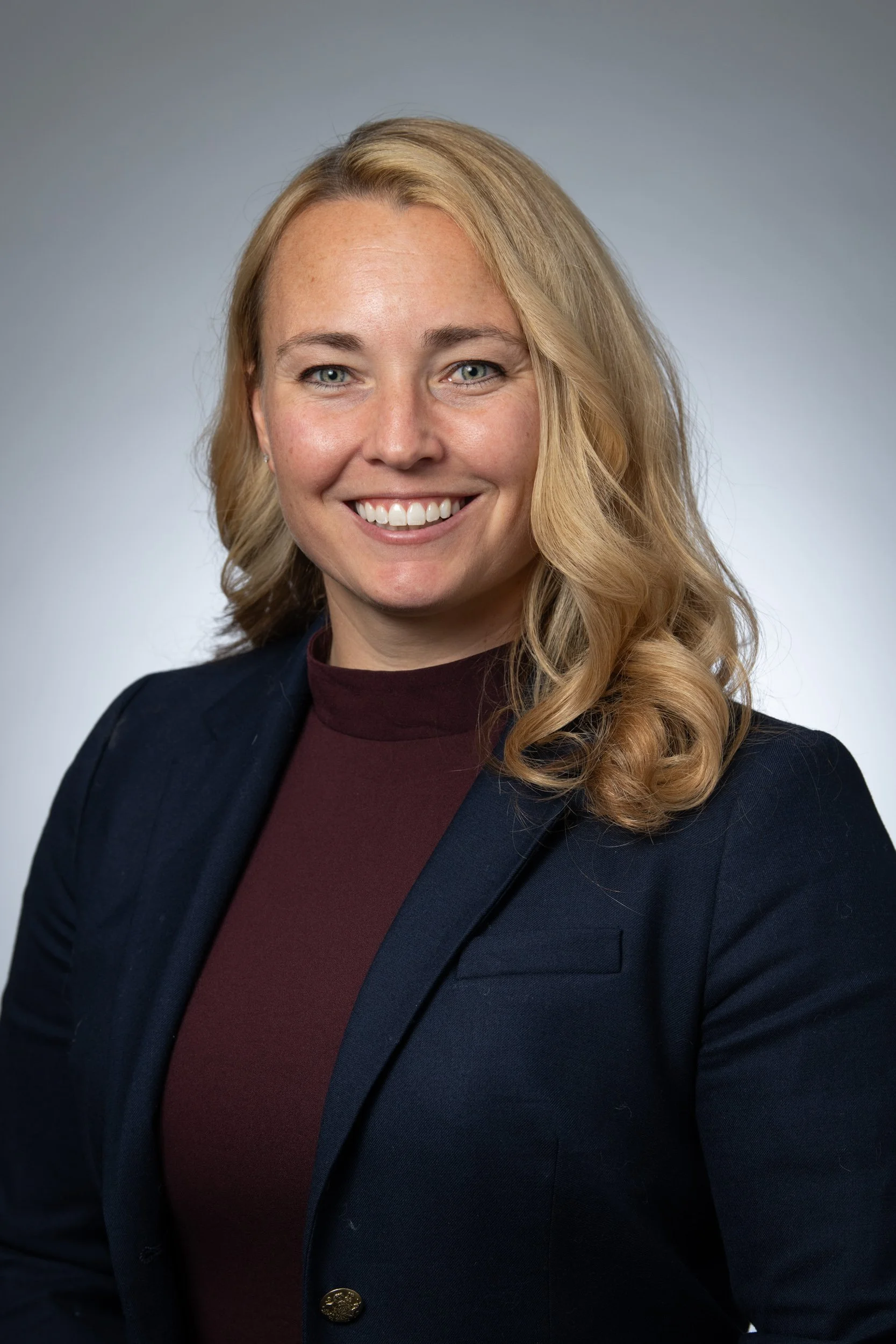Professional woman with blonde curly hair, blue eyes, wearing a dark blazer and maroon top, smiling against a plain grey background.