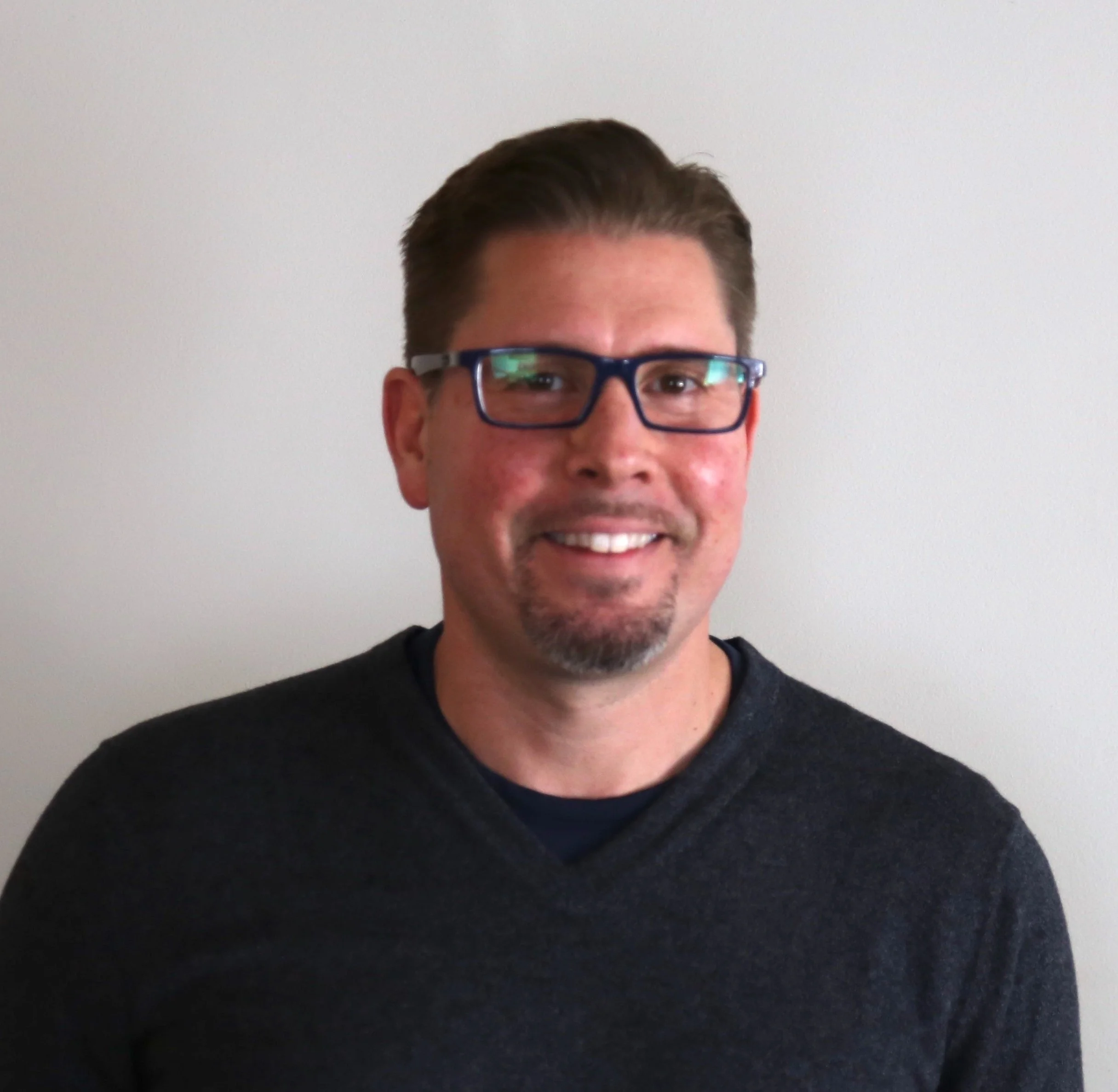 A man with short dark hair, wearing glasses and a dark shirt, smiling in front of a plain, light-colored wall.