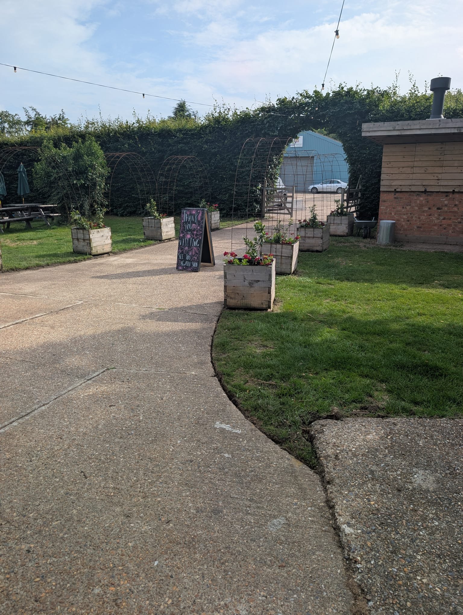 A sidewalk leading to an outdoor area with potted flowers and a blackboard sign reading 'Thank you for visiting, see you again soon.' in front of a small, outdoor patio with string lights overhead.