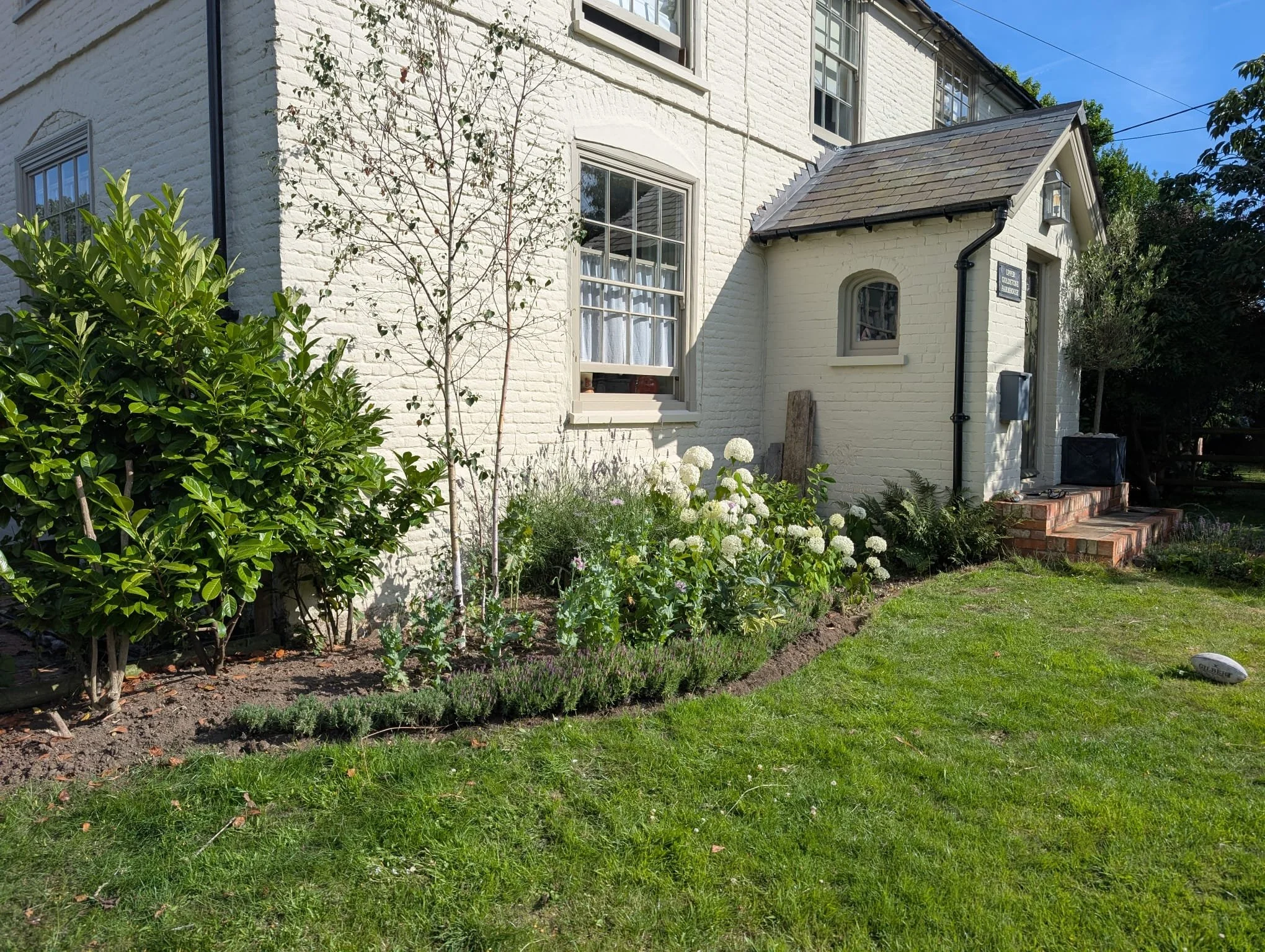 White brick house with a small garden in front, featuring various plants and flowers, including a small tree and shrubbery, on a sunny day.