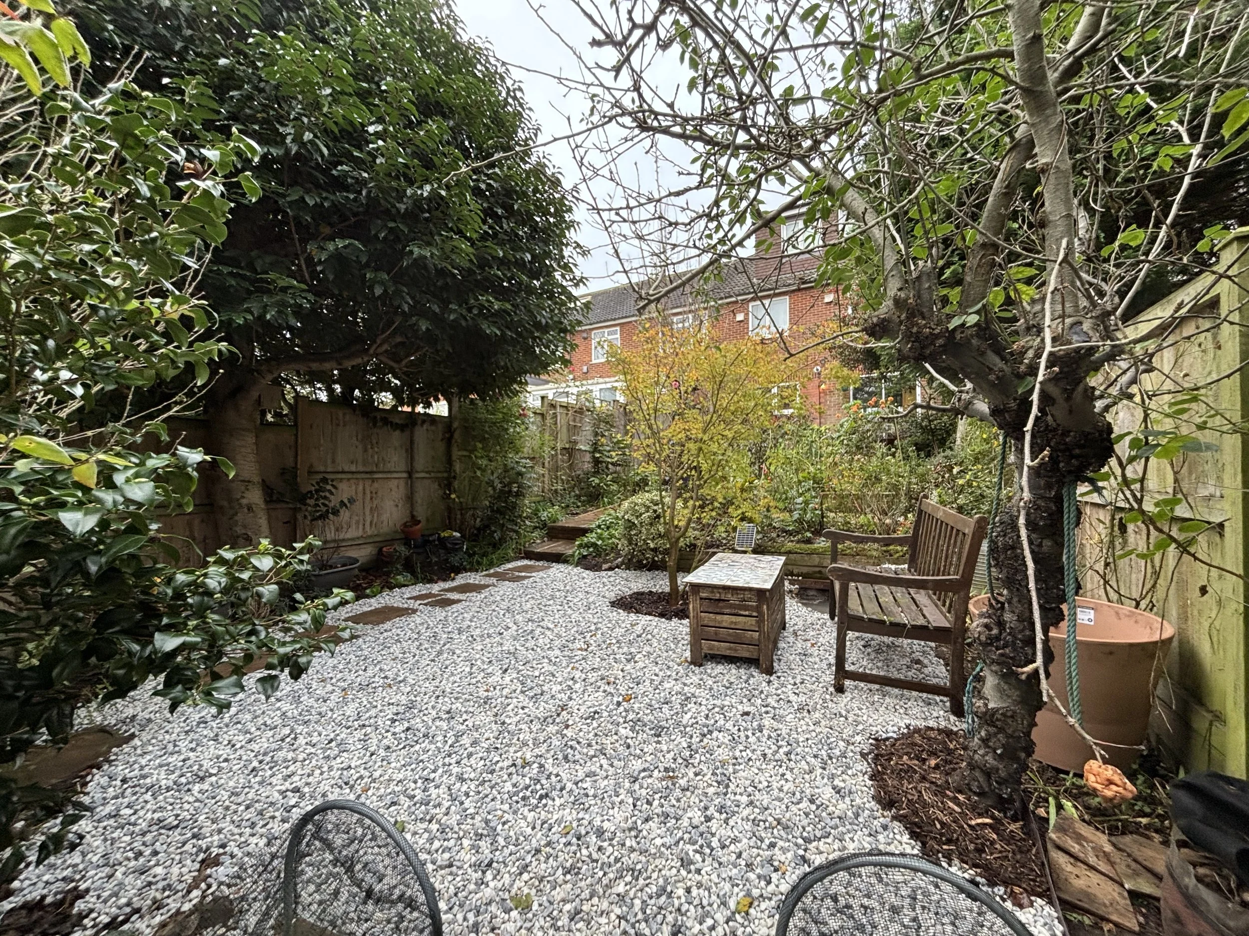 A cozy backyard garden with a gravel pathway, lush green trees, a small yellow tree, a wooden bench, and an orange flower pot. Fencing surrounds the garden with a brick house visible in the background.