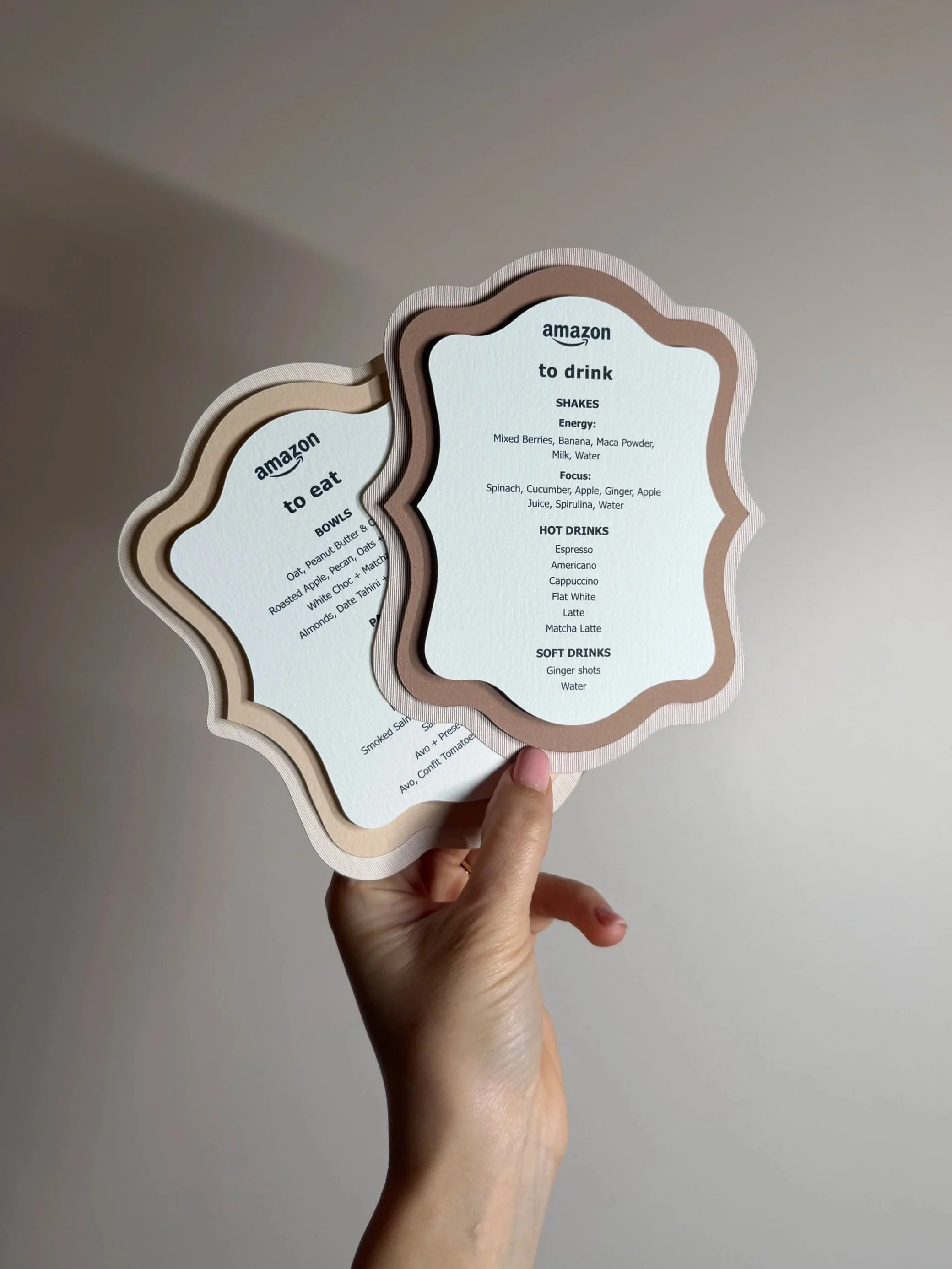 Two layered menus with scalloped edges, held in a person's hand, displaying options for drinks, food, and bowls from Amazon.