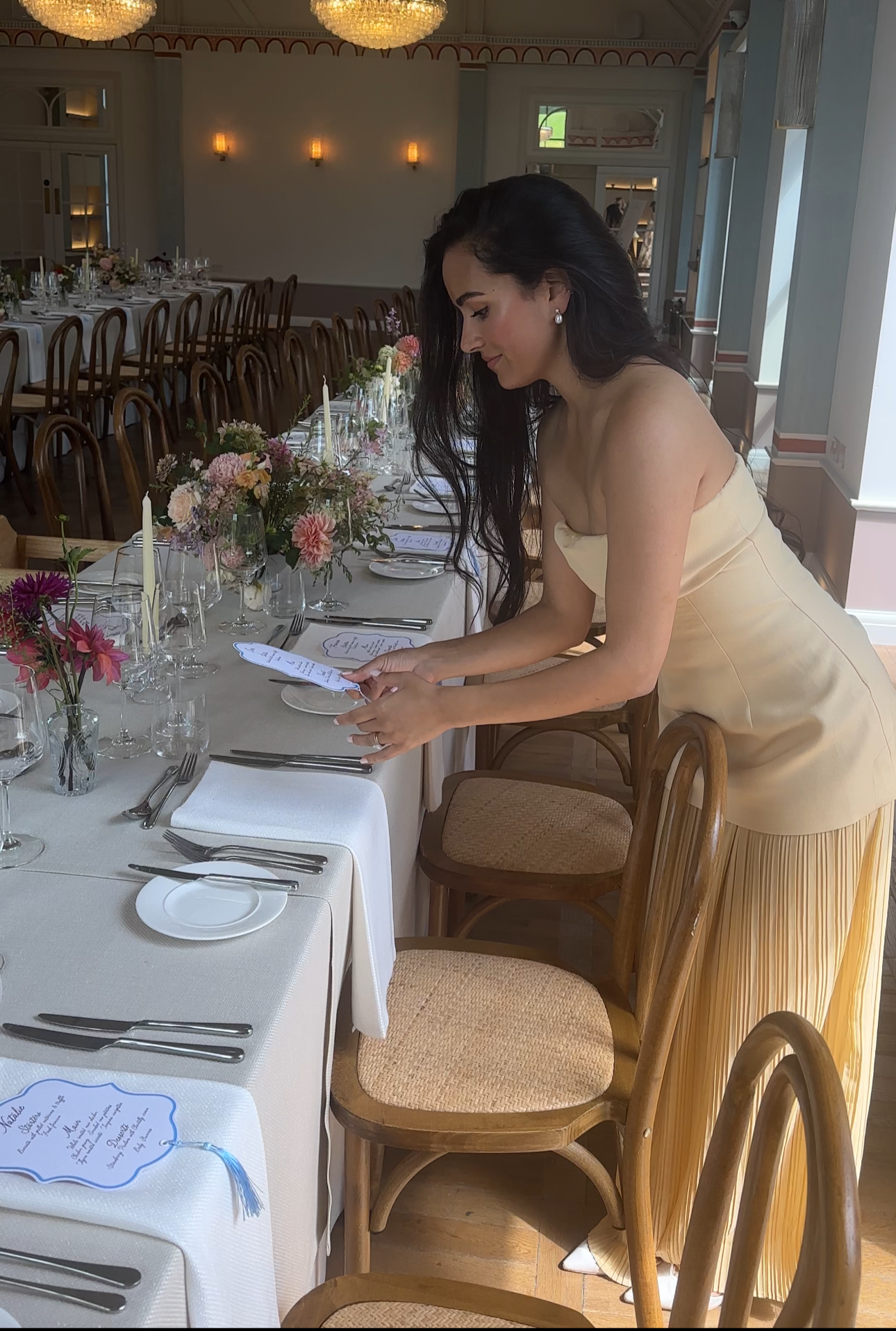 A woman in a strapless, cream-colored dress with a pleated skirt setting a dinner table decorated with pink, purple, and white flowers, and holding a printed event menu.