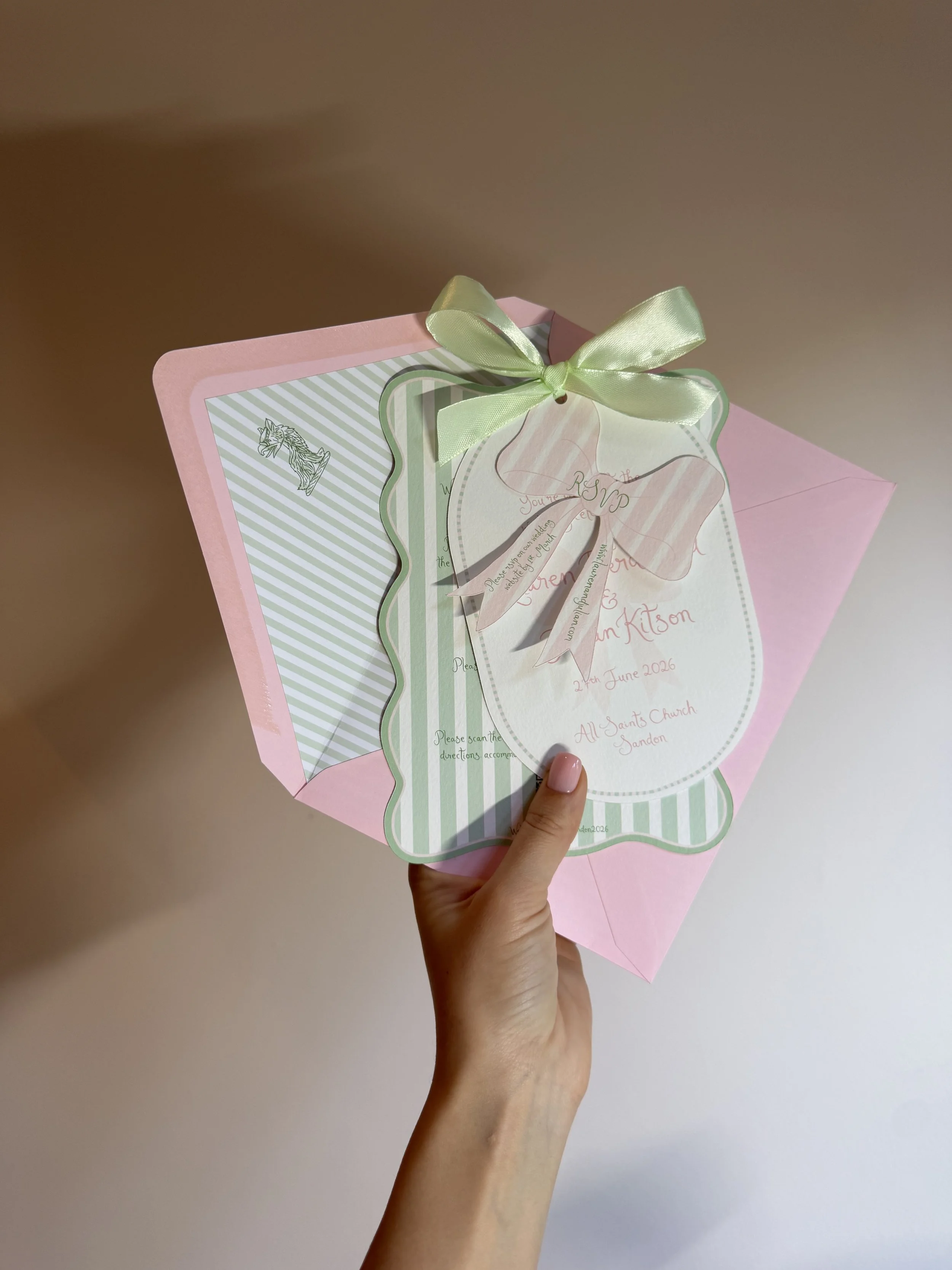 Pink invitation card with green and white striped border, attached to a pink envelope, decorated with a green satin bow, for a wedding event on June 2, 2026, at All Saints Church in Jarden.