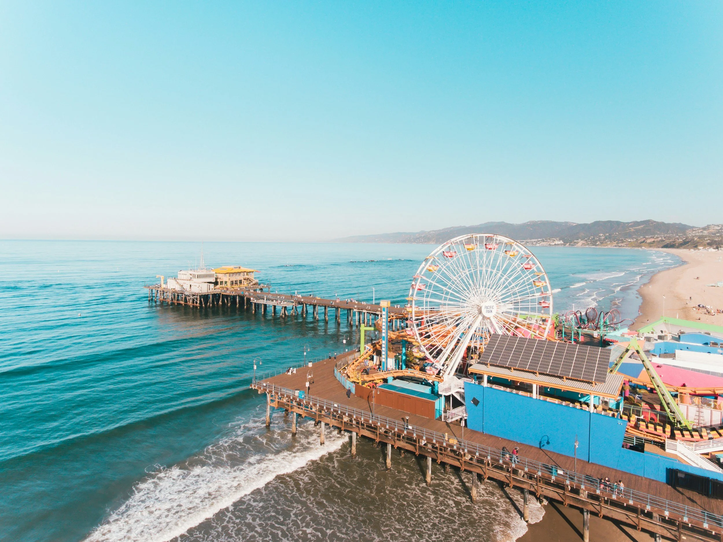 Aerial view of Santa Monia Pier with a Ferris wheel, roller coaster, and a pier extending into the ocean, with sandy beach and hills in the background.