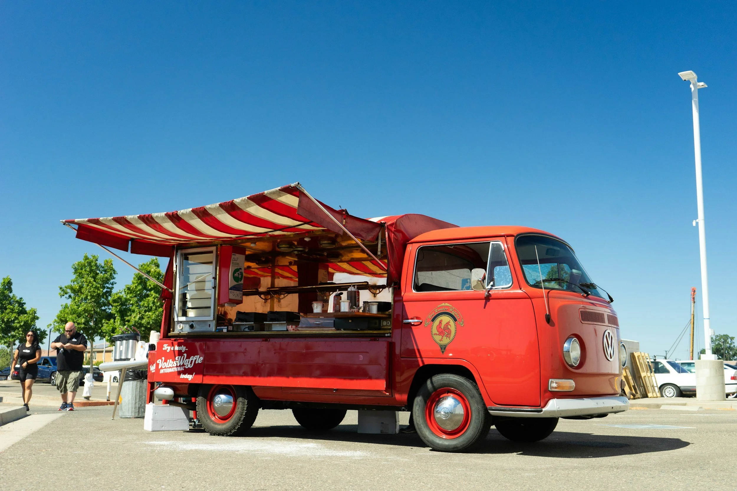 Red vintage Volkswagen van converted into a food truck with a red and white striped awning, parked in a lot with a few people walking nearby, sunny day with clear blue sky and green trees in the background.
