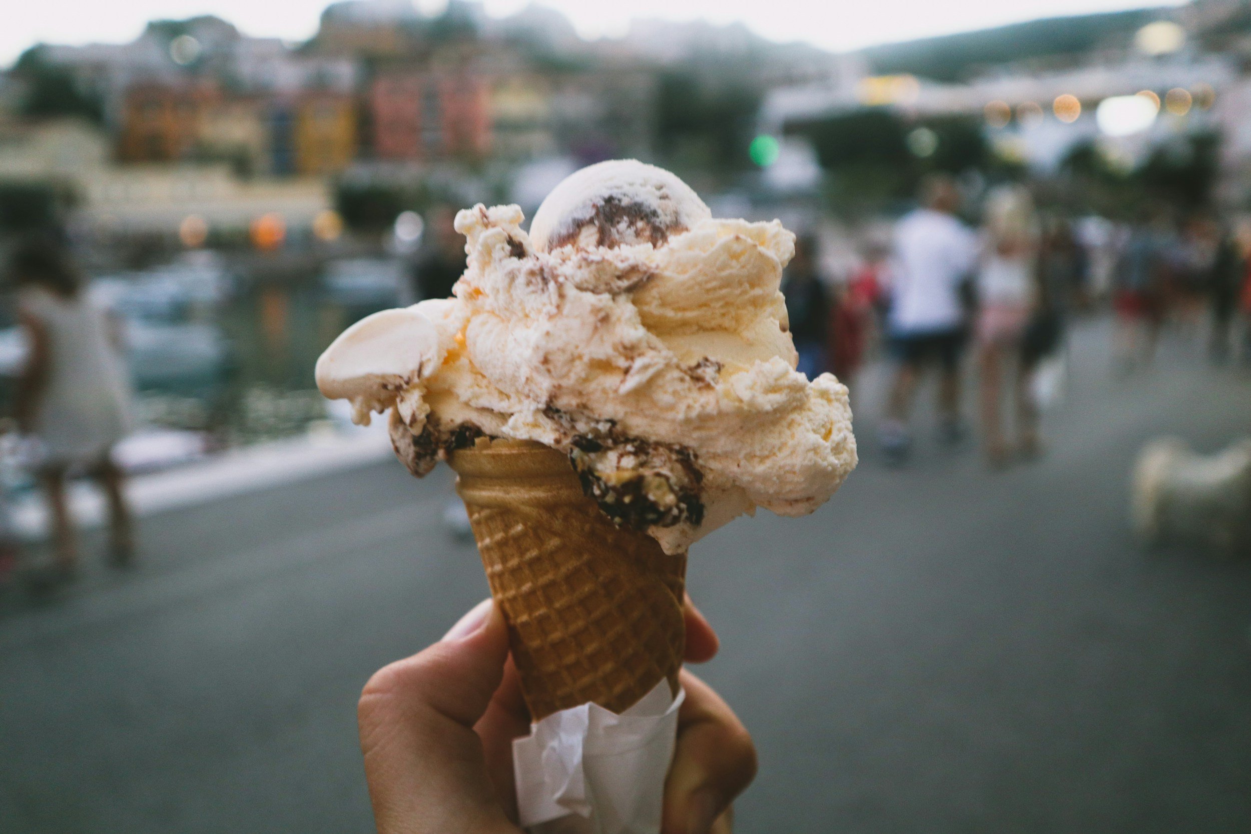 Close-up of a scoop of ice cream in a waffle cone, with a blurry background of people near a waterfront with boats and buildings.