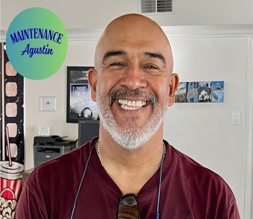 A smiling man with a gray beard and sunglasses hanging from his shirt standing in a room with a TV and pictures on the wall, featuring a green circle sticker in the top left corner that reads "Maintenance Agustin".