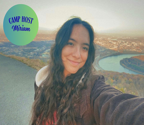 Young woman taking a selfie outdoors with a valley and river in the background, during sunset. There is a green and blue camp sign with text reading 'Camp Host Miriam'.