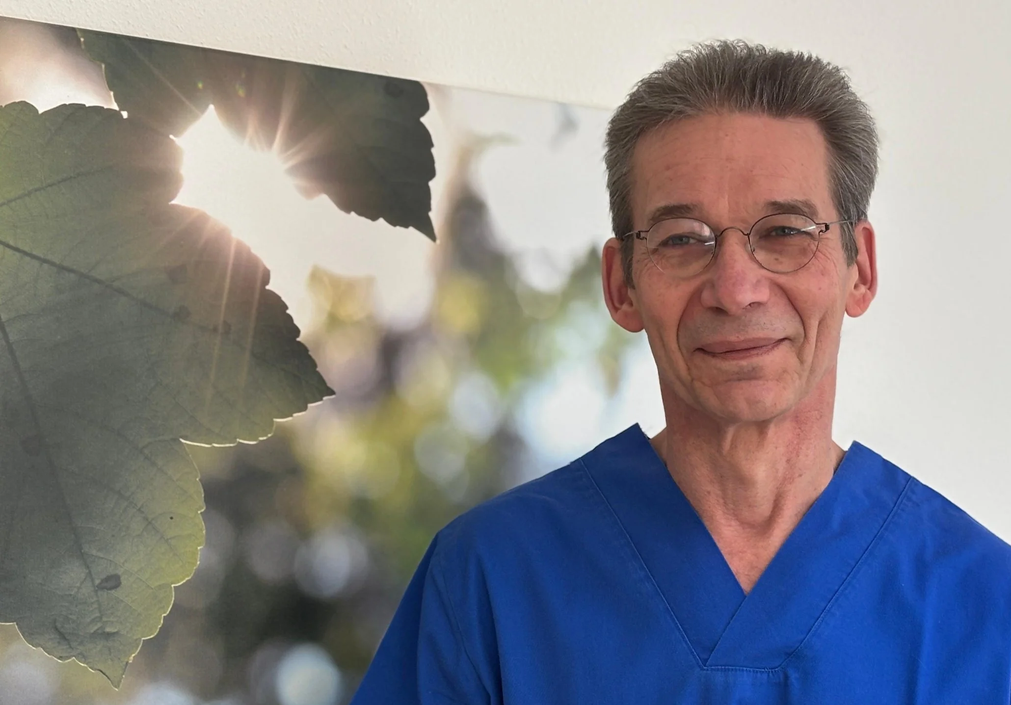 Ein Mann in blauer medizinischer Kleidung steht vor einem Hintergrundbild mit Blättern und Sonnenlicht.
