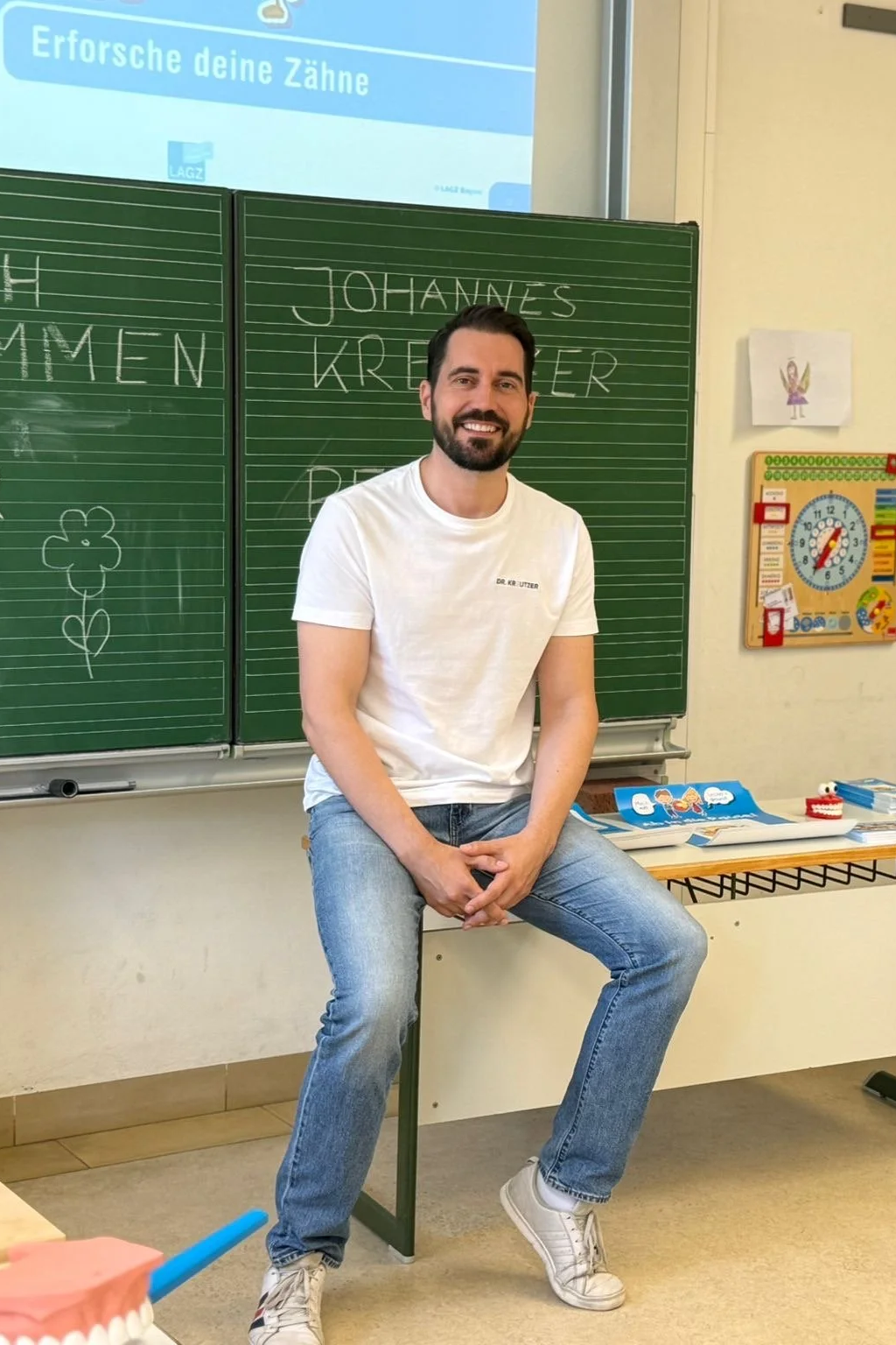 Ein Mann sitzt vor einer Schul-Tafel in einem Klassenzimmer, lächelt und trägt ein weißes T-Shirt mit Aufdruck, daneben Lehrmaterialien und eine Tafel.