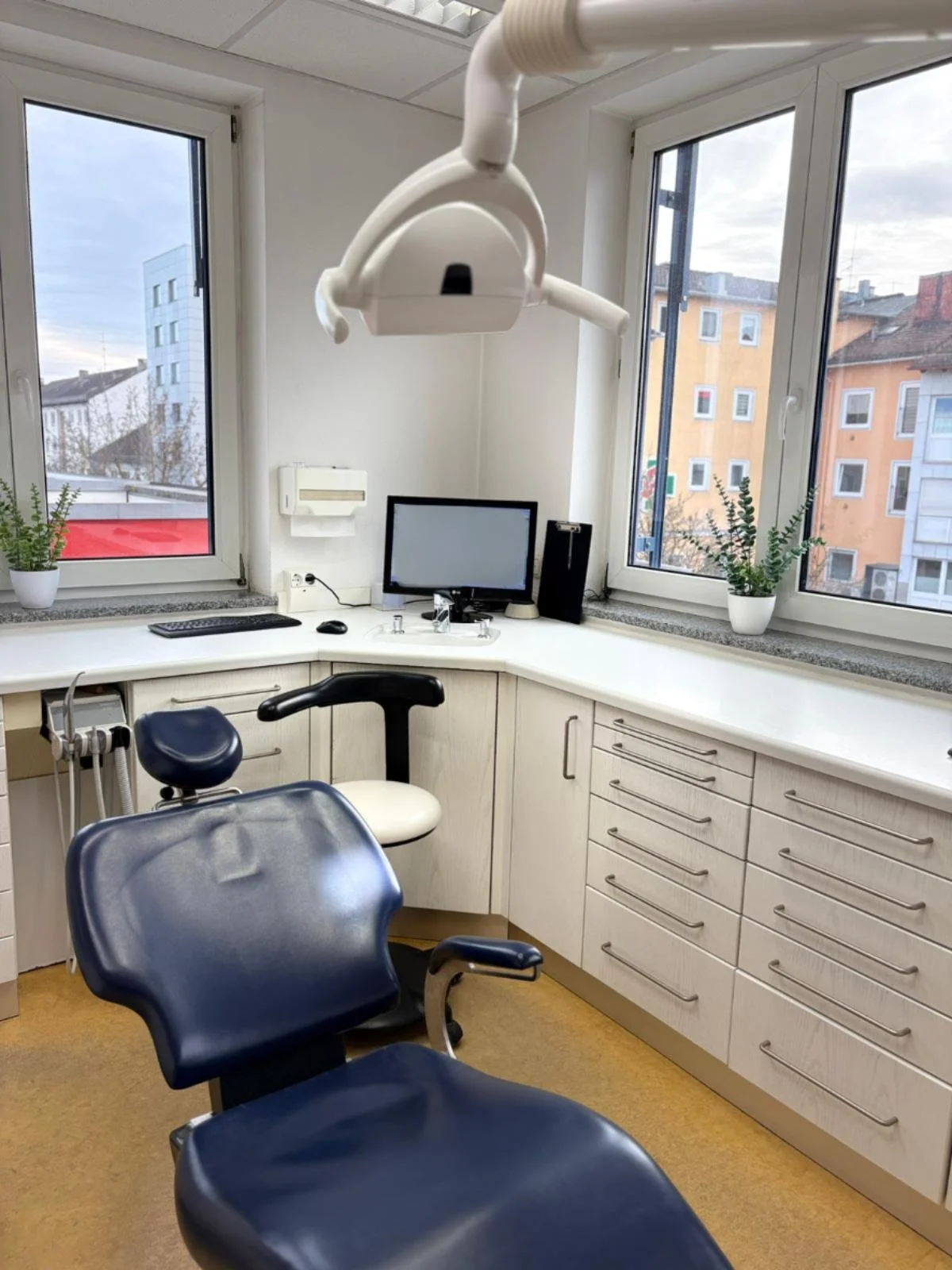 Behandlungsraum mit zahnärztlichem Stuhl, Arbeitslampe, Computer, Fenstern mit Stadtblick, Pflanzen und medizinischer Ausstattung.