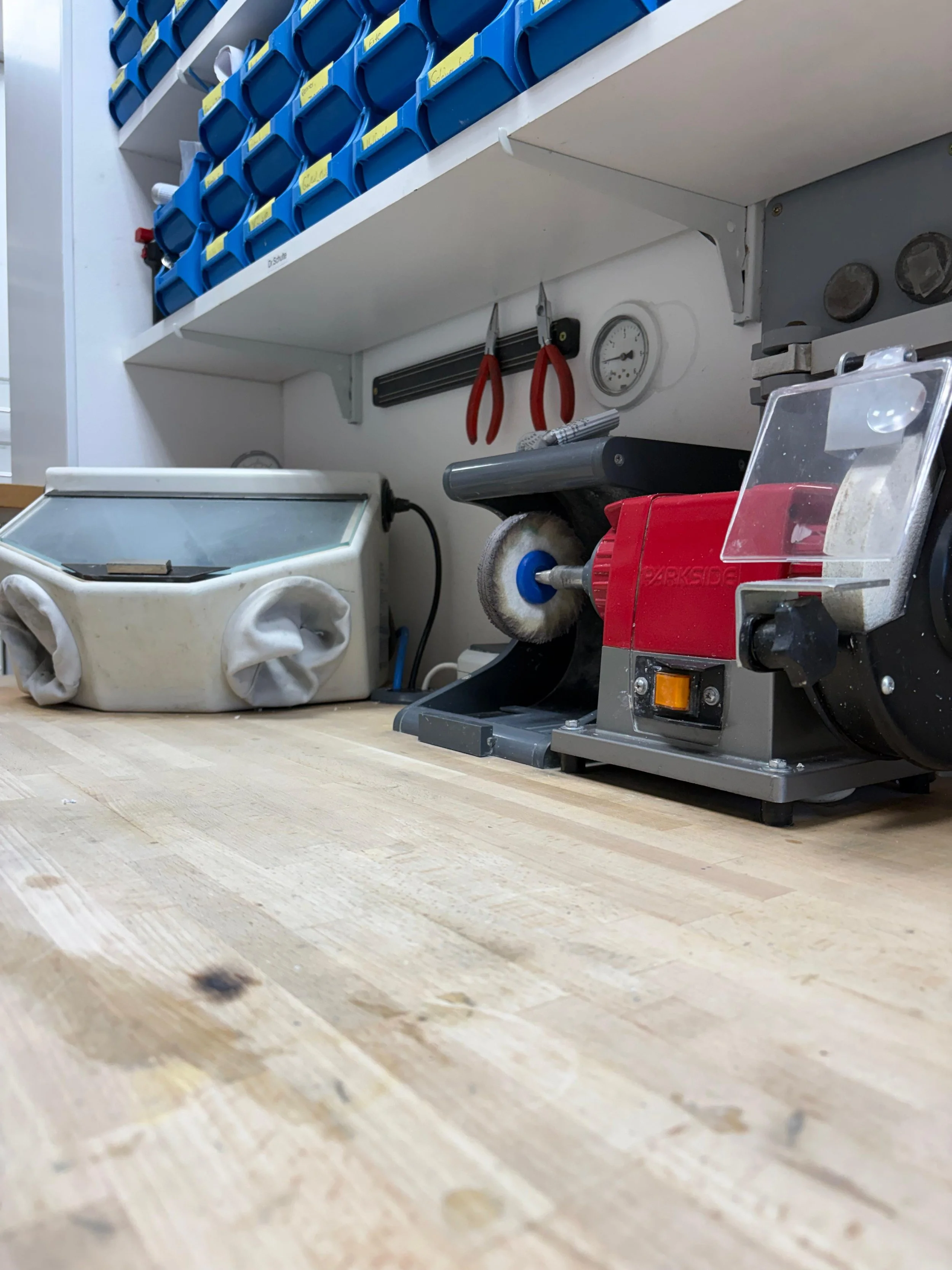 Werkzeug und Materialien auf einer Holzarbeitsplatte in einem Reparatur- oder Werkstattbereich, darunter eine rote Schleifmaschine, Pinsel, Schrauben und ein Druckmesser.