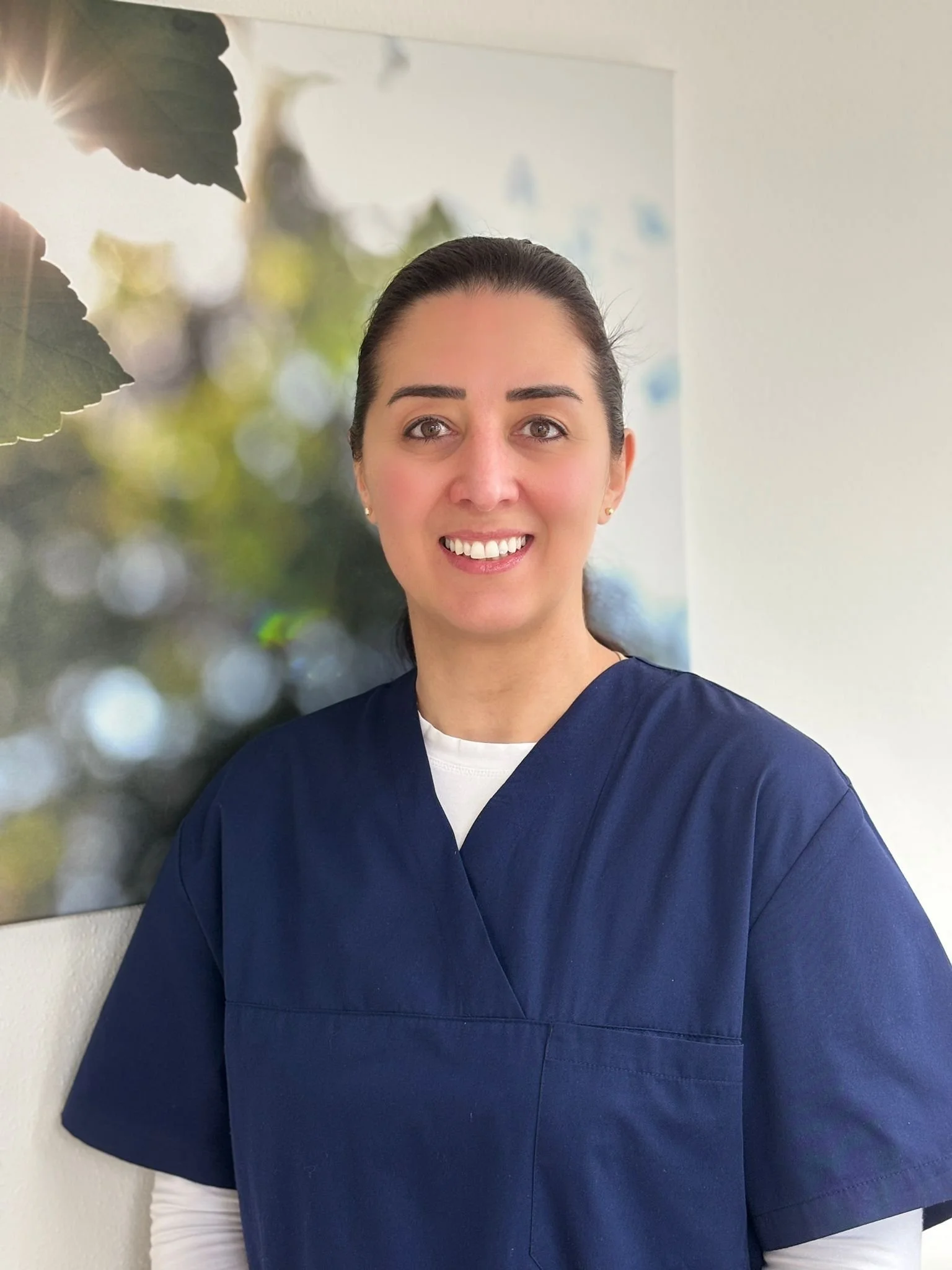 Eine lächelnde Frau in blauer Krankenhausschürze steht vor einem Heilungsbild an der Wand.