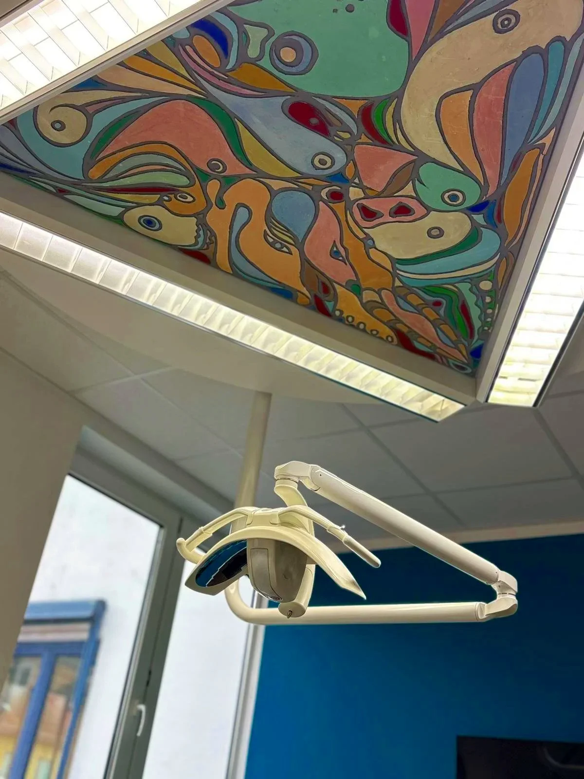 Deckenleuchte mit buntem abstraktem Kunstwerk im Hintergrund, wahrscheinlich in einem medizinischen Raum, da eine zahnärztliche Lampe sichtbar ist.