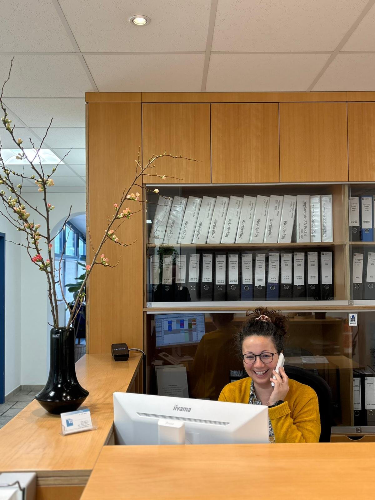 Eine Frau sitzt an einem Empfangstresen, lächelt und spricht am Telefon. Im Hintergrund sind Aktenregale, eine große Vase mit Zweigen und eine Empfangsanzeige sichtbar.