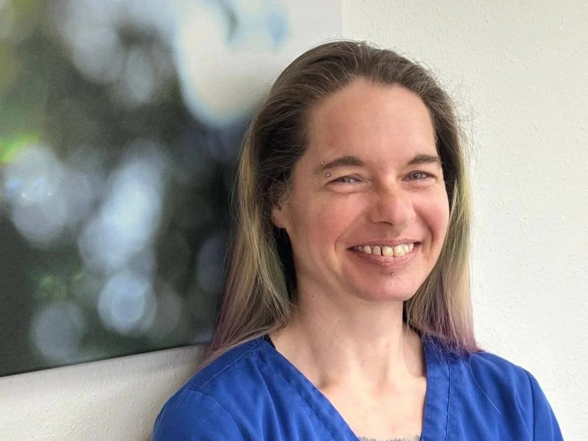 Eine Frau mit langen braunen Haaren, die ein Lächeln zeigt, trägt eine blaue Schürze und steht vor einem weißen Wand. Hinter ihr ist ein unscharfes Bild mit Kreiseffekt.