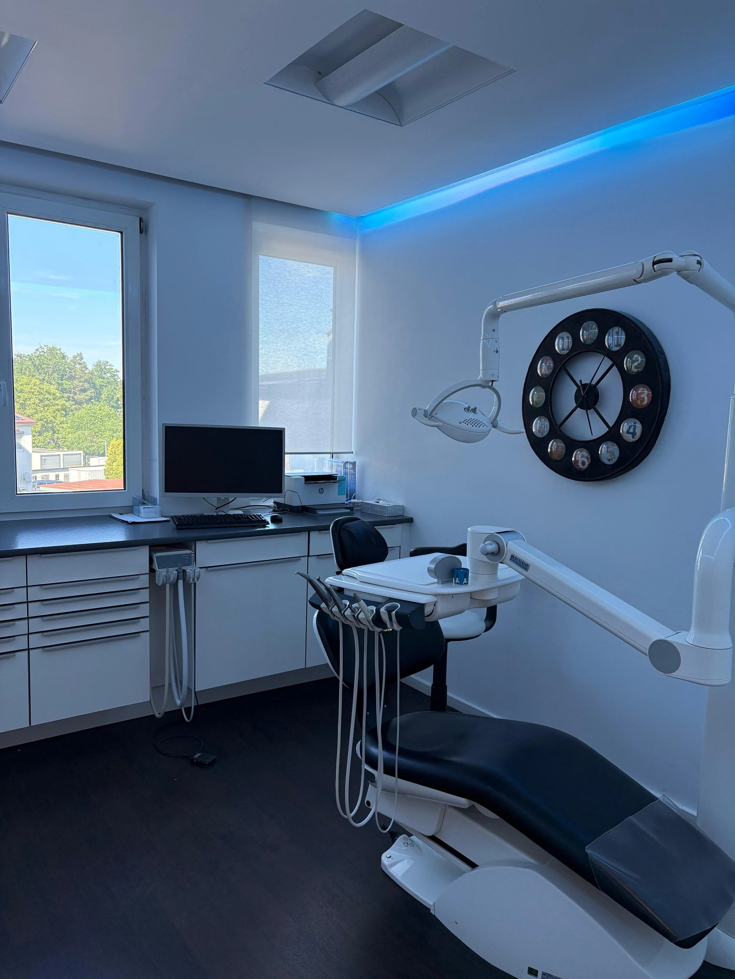 Ein Behandlungsraum in einer Zahnarztpraxis mit einer Liege, einem Computerbildschirm, medizinischen Geräten und einem großen Wandkreisel. Das Zimmer ist hell erleuchtet und hat große Fenster mit Blick nach draußen.