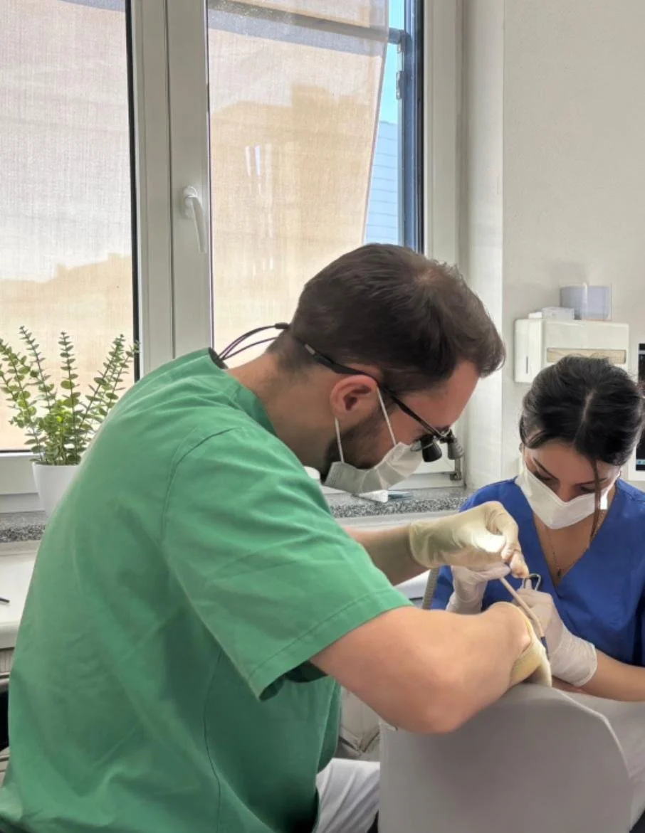 Zwei medizinische Fachkräfte, ein Mann und eine Frau, führen eine medizinische Behandlung in einem Behandlungsraum durch, beide tragen Masken und Handschuhe.