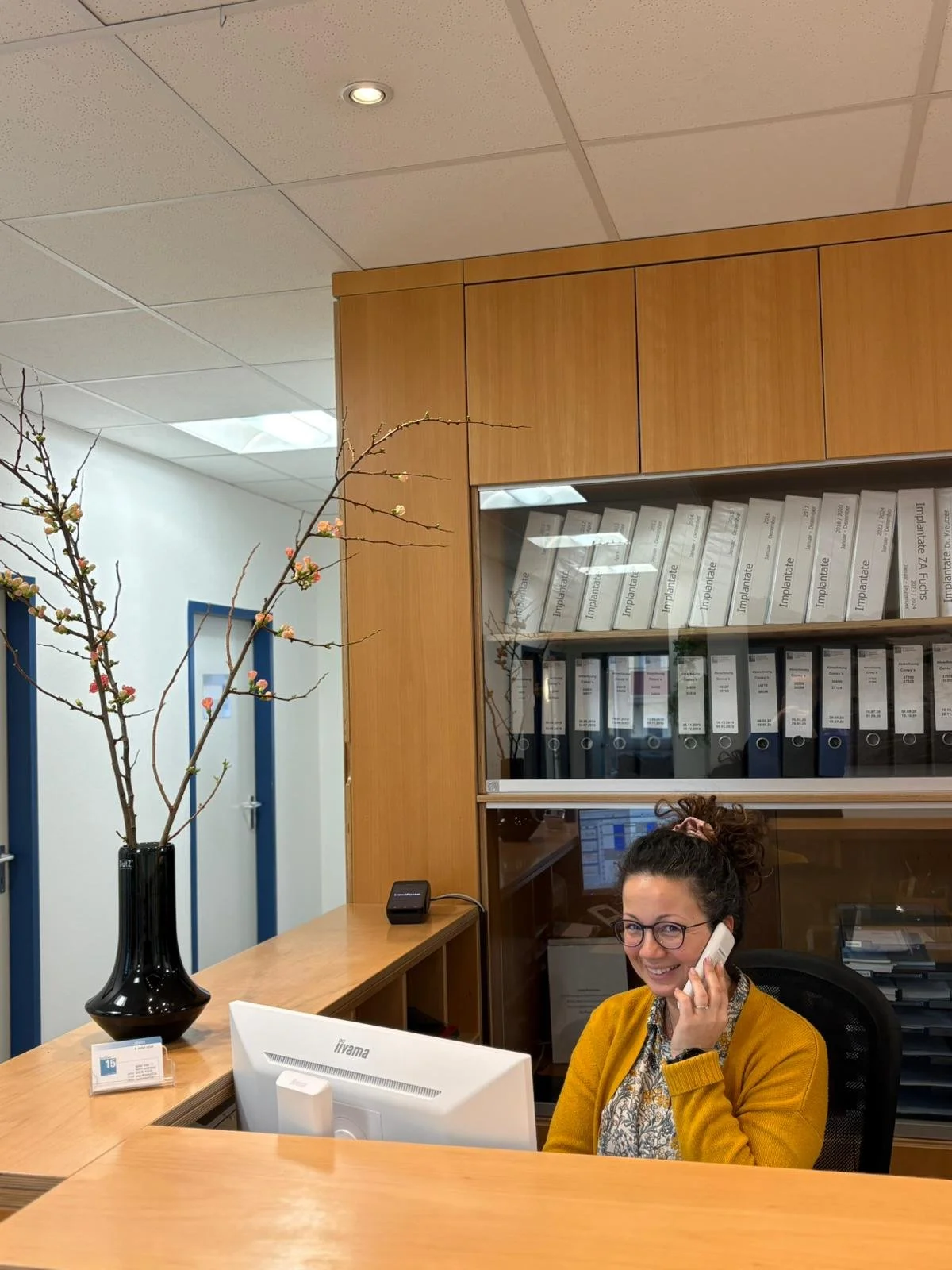 Eine Frau sitzt an einem Empfangstresen, trägt eine gelbe Jacke und spricht am Telefon. Im Hintergrund ein Regal mit Aktenordnern und eine Vase mit Zweigen auf dem Tisch.