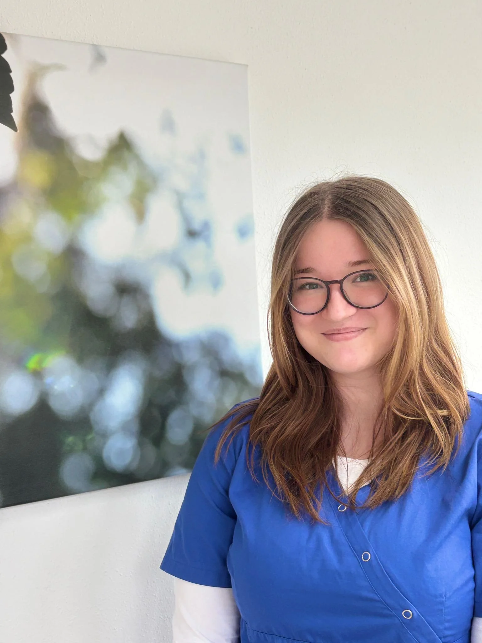 Junge Frau mit brünettem Haar und Brille, die in blauer Arbeitskleidung vor einer weißen Wand steht, im Hintergrund ein verschwommenes Wandbild.