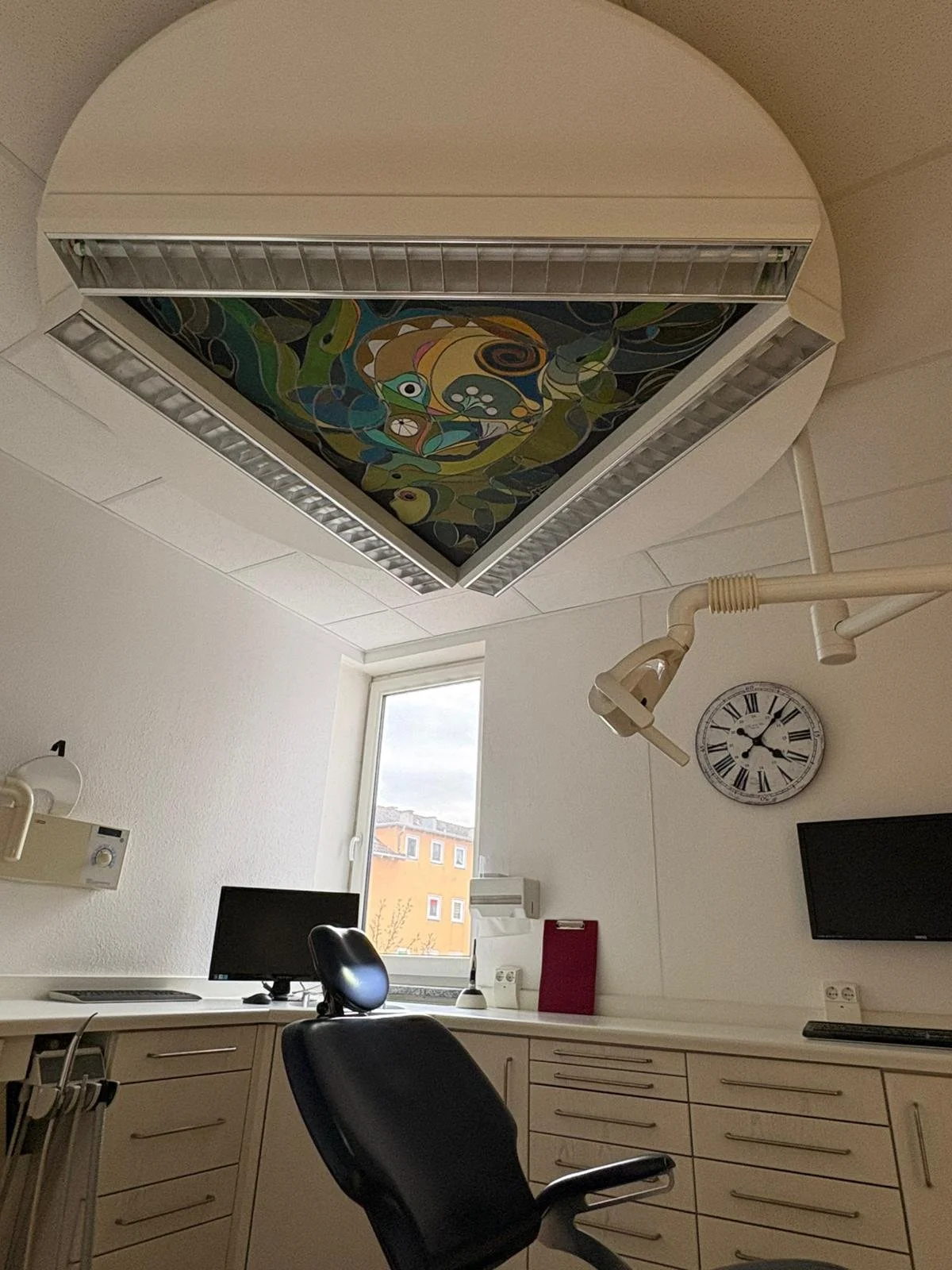 Blick in ein medizinisches Behandlungszimmer mit geschwungenem weißen Deckenstrahler, einem caracteristischen Decke Musters, einem Computerbildschirm, einer Uhr und einem Behandlungsarm. Der Raum hat eine Fenster mit Blick auf eine mehrstöckige gelbe und orange Häuser in der Stadt.