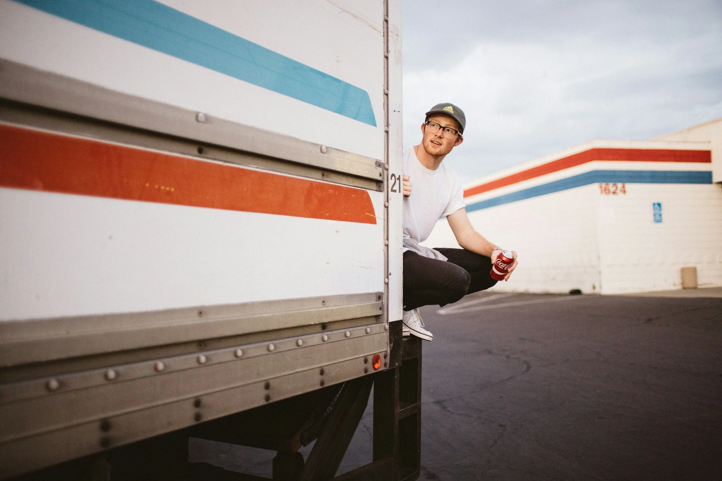 A young man with glasses, a cap, and casual clothes crouches beside a truck, holding a Coca-Cola can and looking to the side, in an outdoor parking lot with trailers and a cloudy sky.