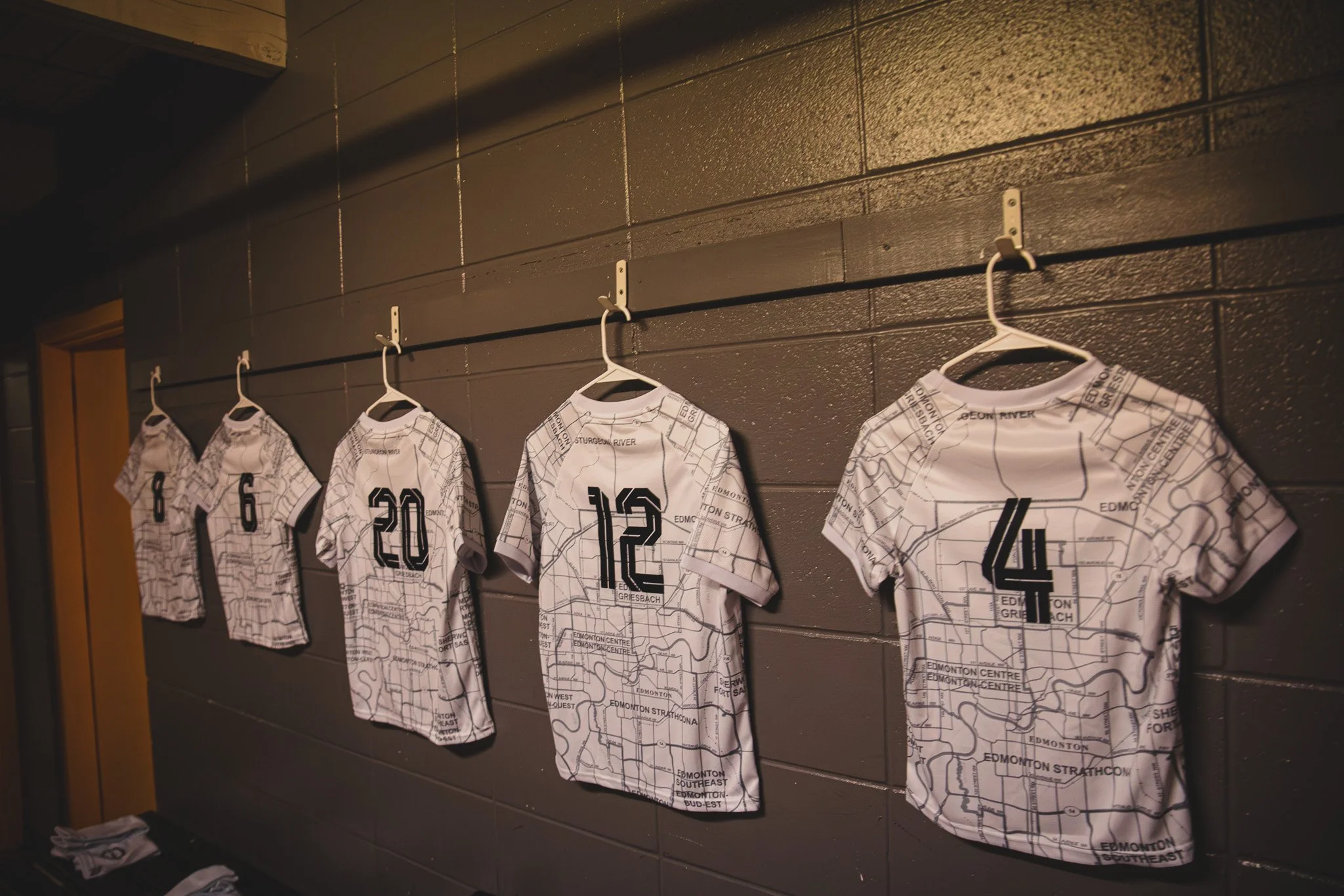 Multiple white sports jerseys with black numbers and a city map design, hanging on hooks against a dark wall.